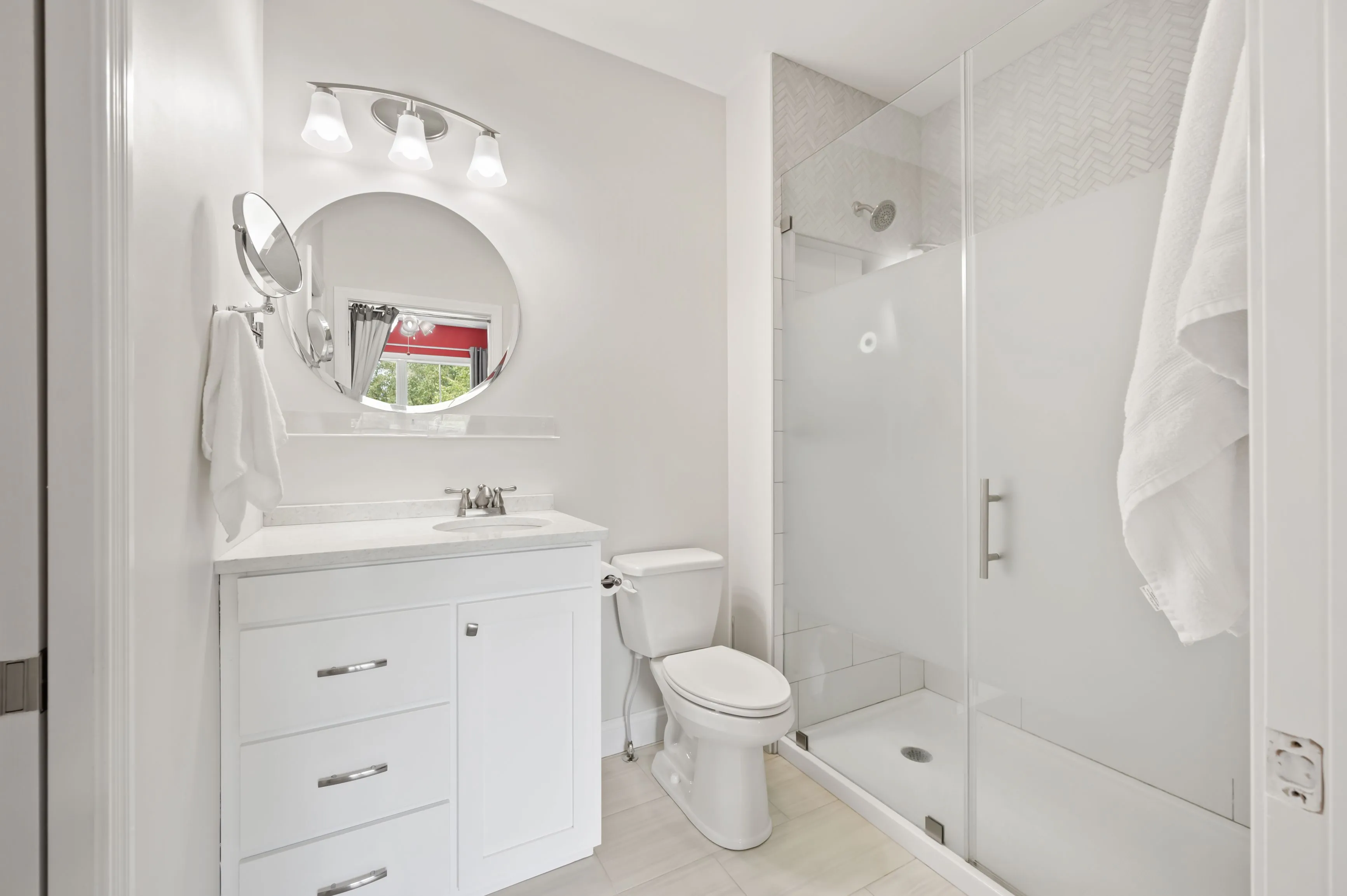 Bright modern bathroom with a white vanity, circular mirror, toilet, and shower area.