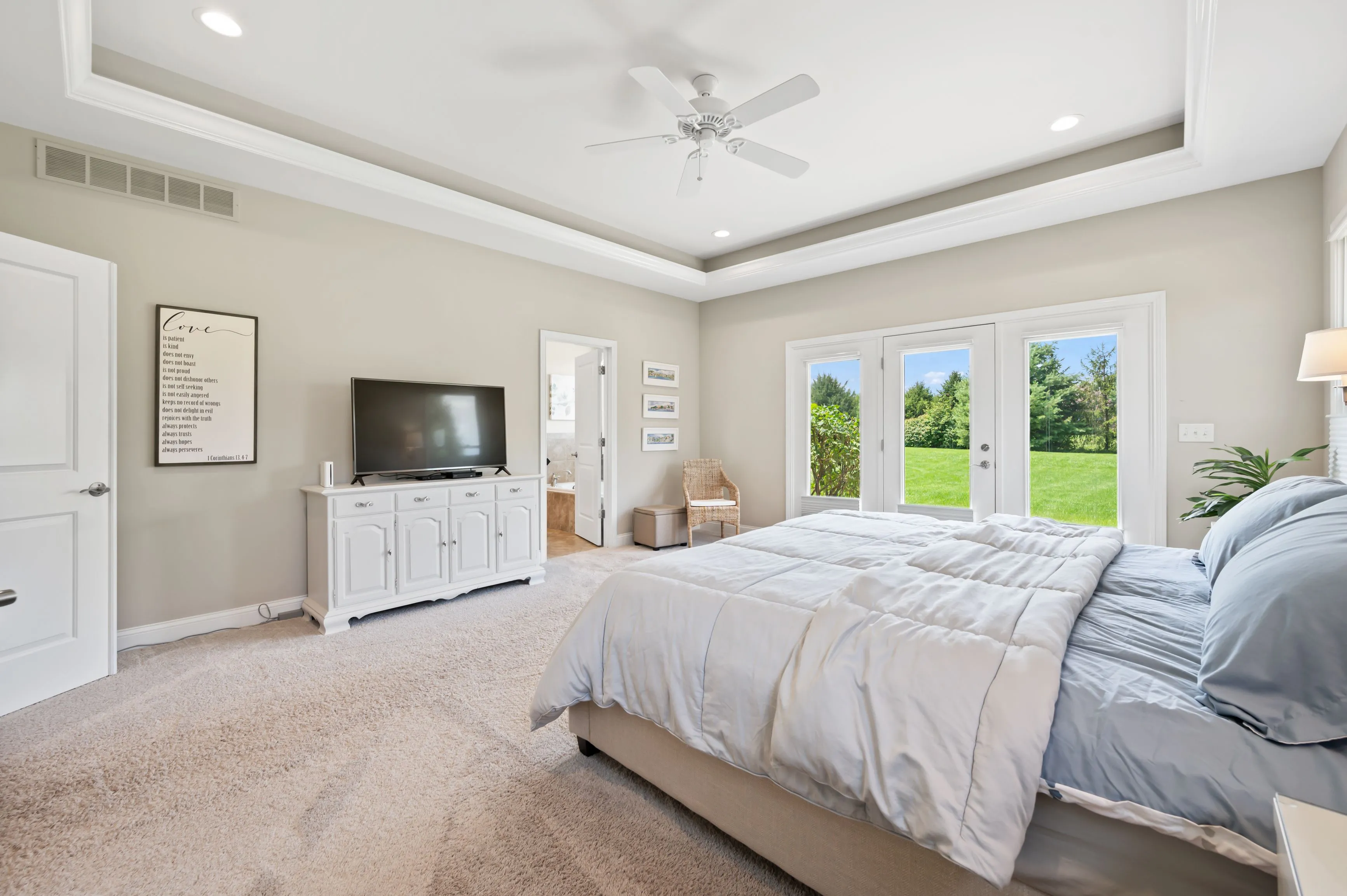 Bright and spacious bedroom with a large bed, white linens, ceiling fan, and a view to the garden through open French doors.