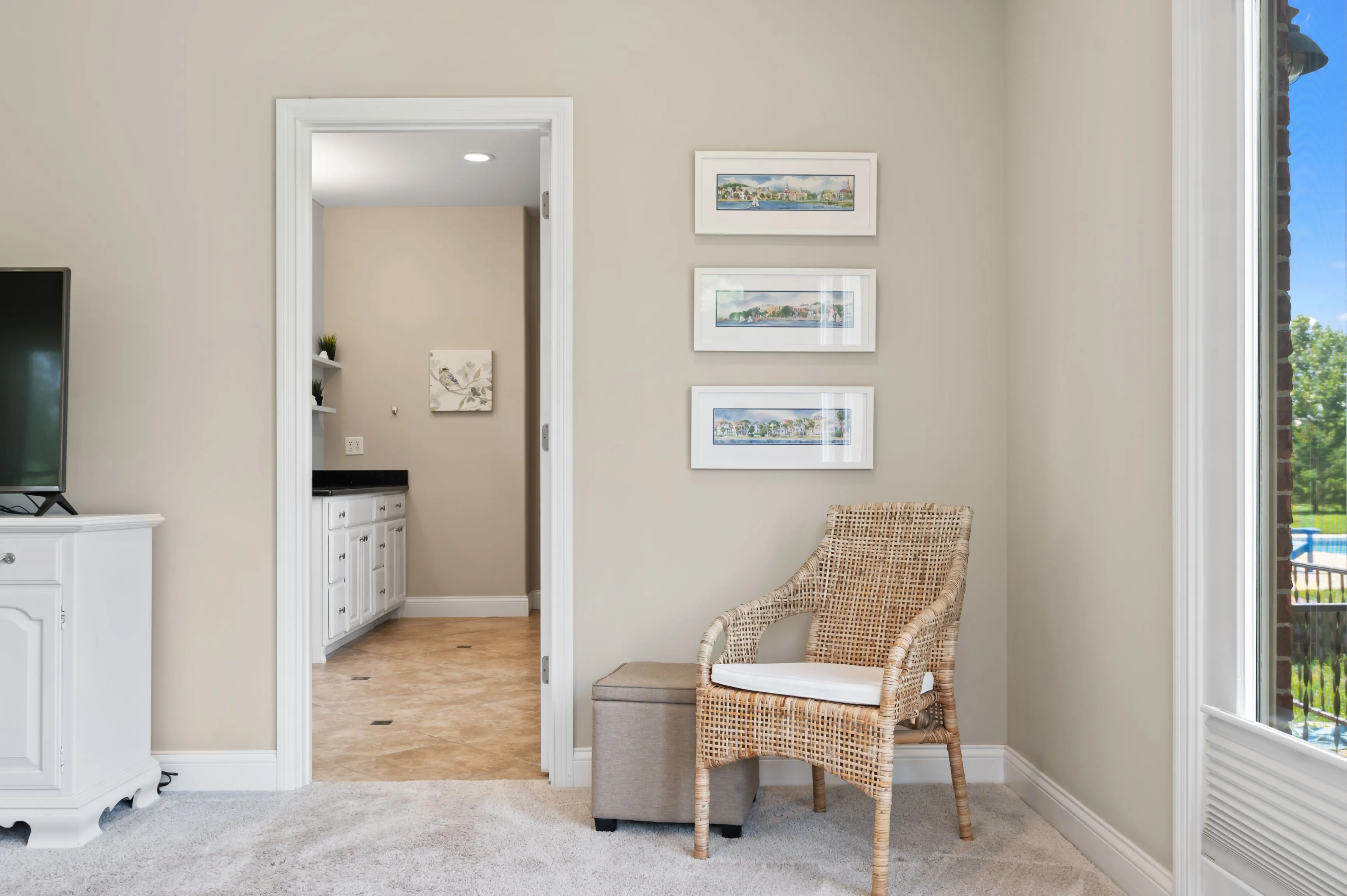A modern hallway with beige walls, featuring a wicker bench, framed pictures on the wall, and an open door leading to another room.