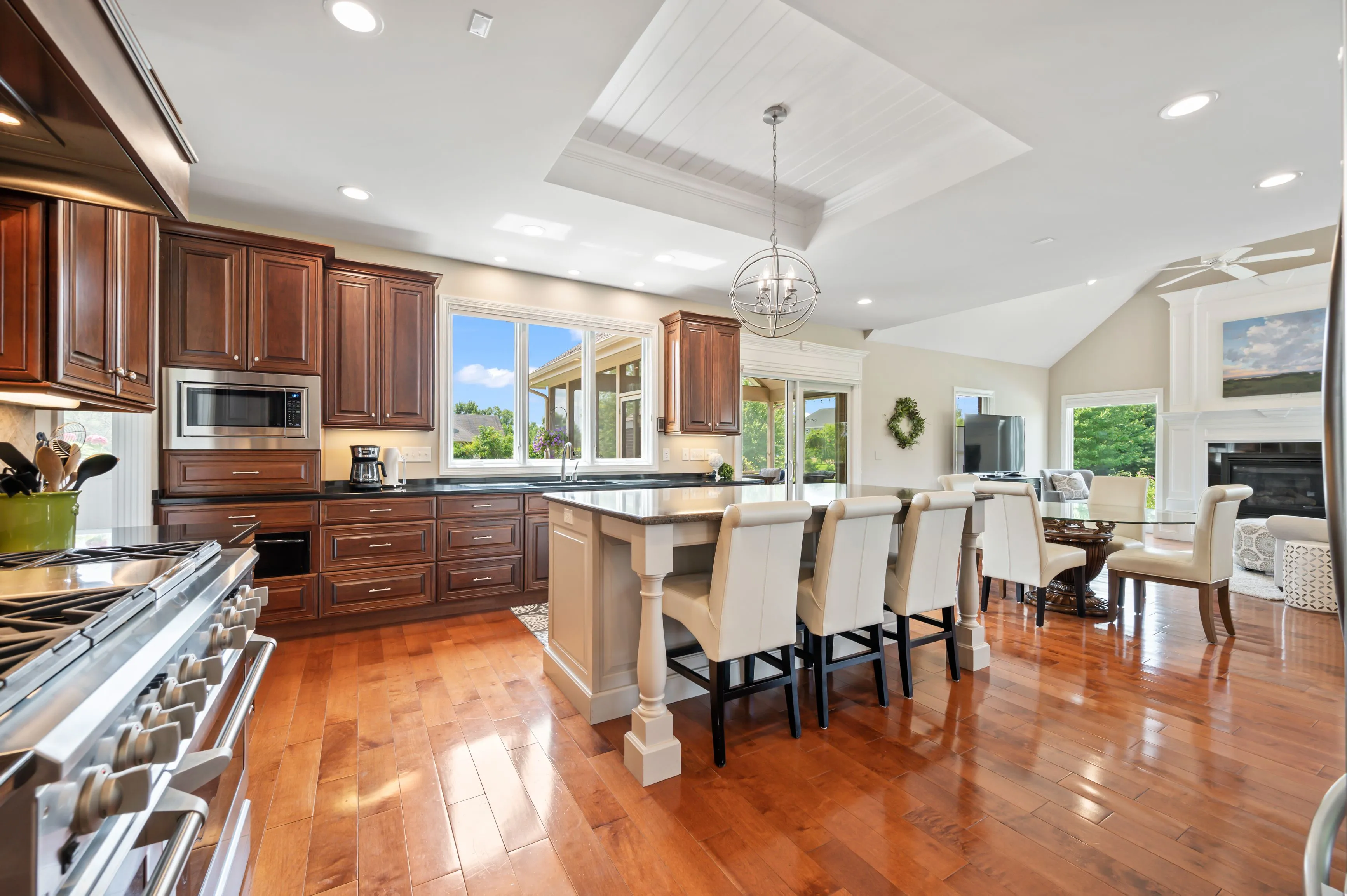 Spacious kitchen with rich wooden cabinetry, stainless steel appliances, a large island with bar stools, and a dining area in the background.