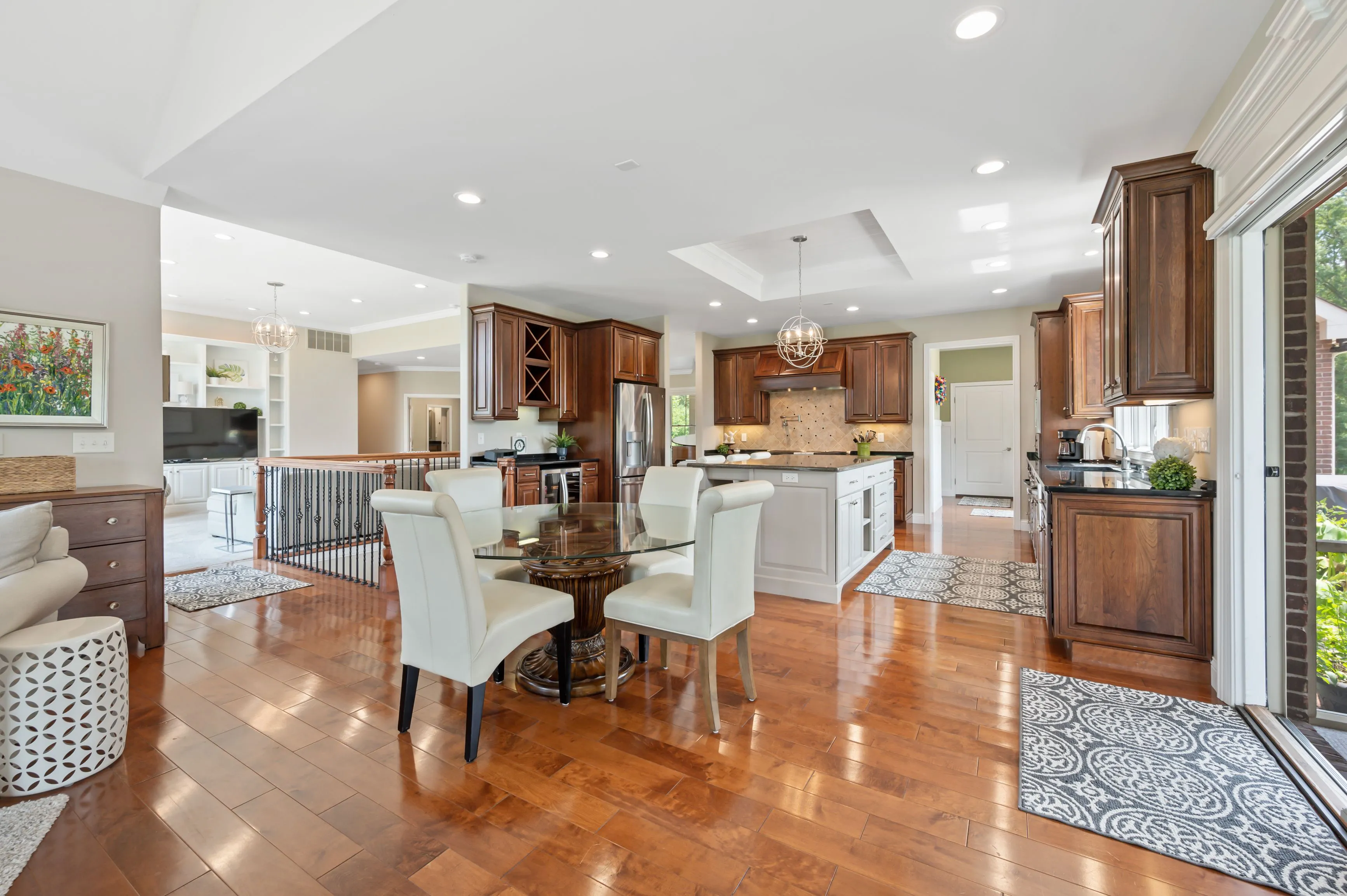 Spacious modern kitchen with polished wooden floors, white dining set, and stainless steel appliances, opening to a living area.