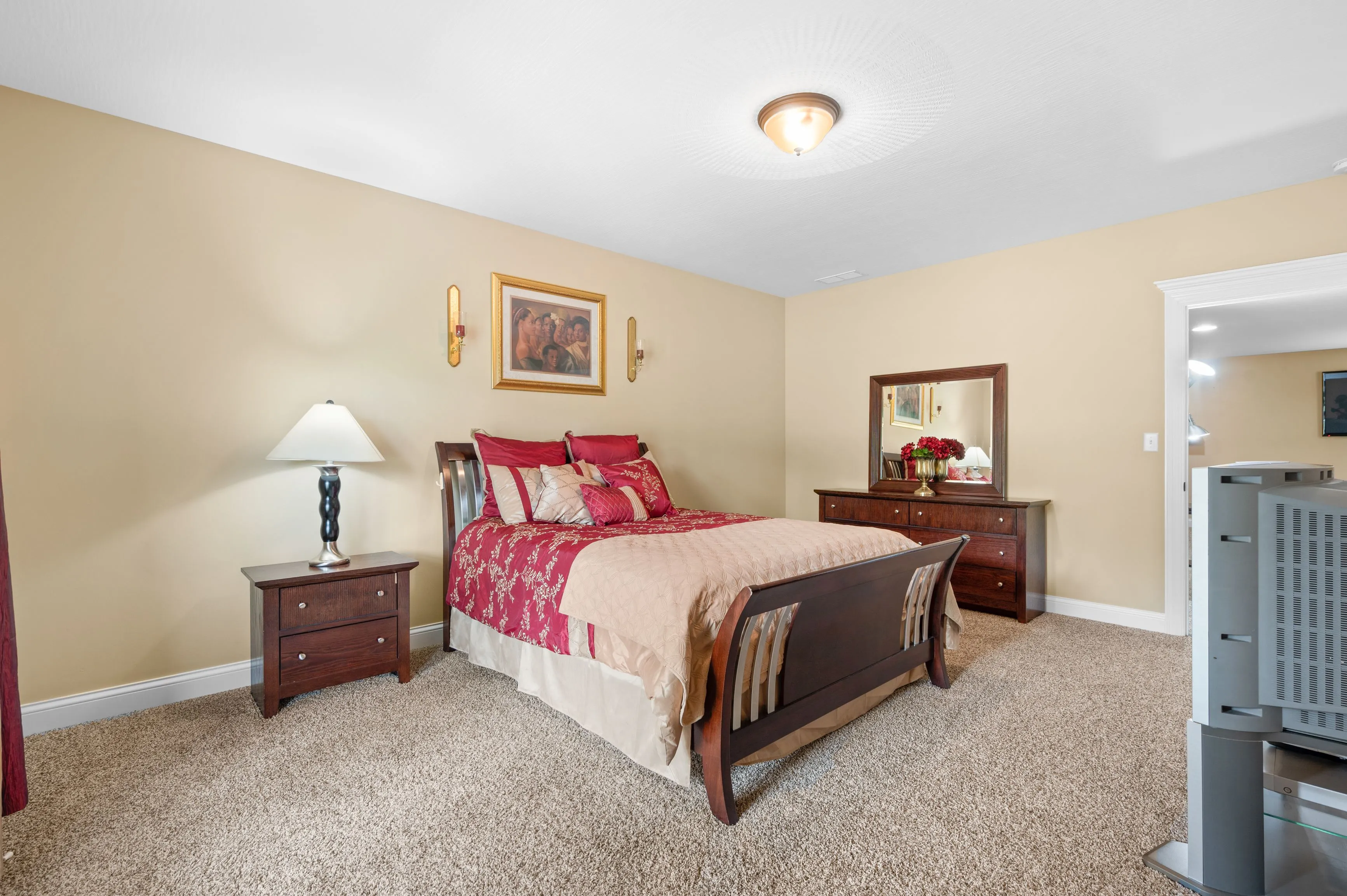 Cozy bedroom interior with a queen-sized bed adorned with red and beige bedding, matching wooden nightstand and dresser, a table lamp, wall art, plush carpeting, and a ceiling light fixture.