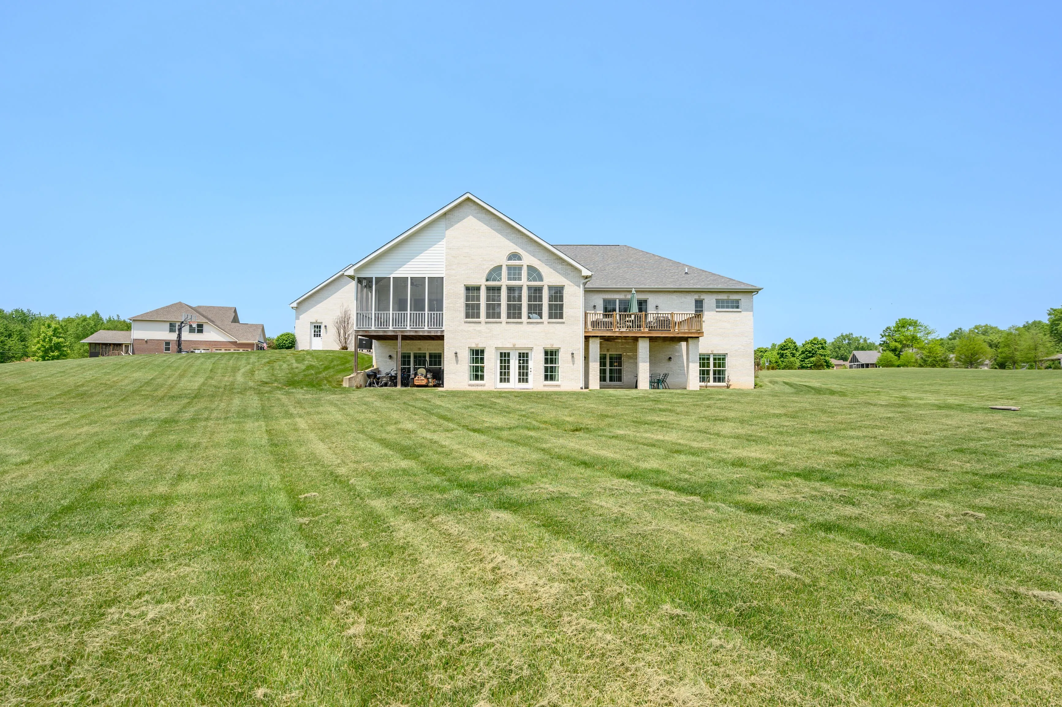 Spacious two-story house with large windows and a wooden deck overlooking an expansive lawn under a clear blue sky.