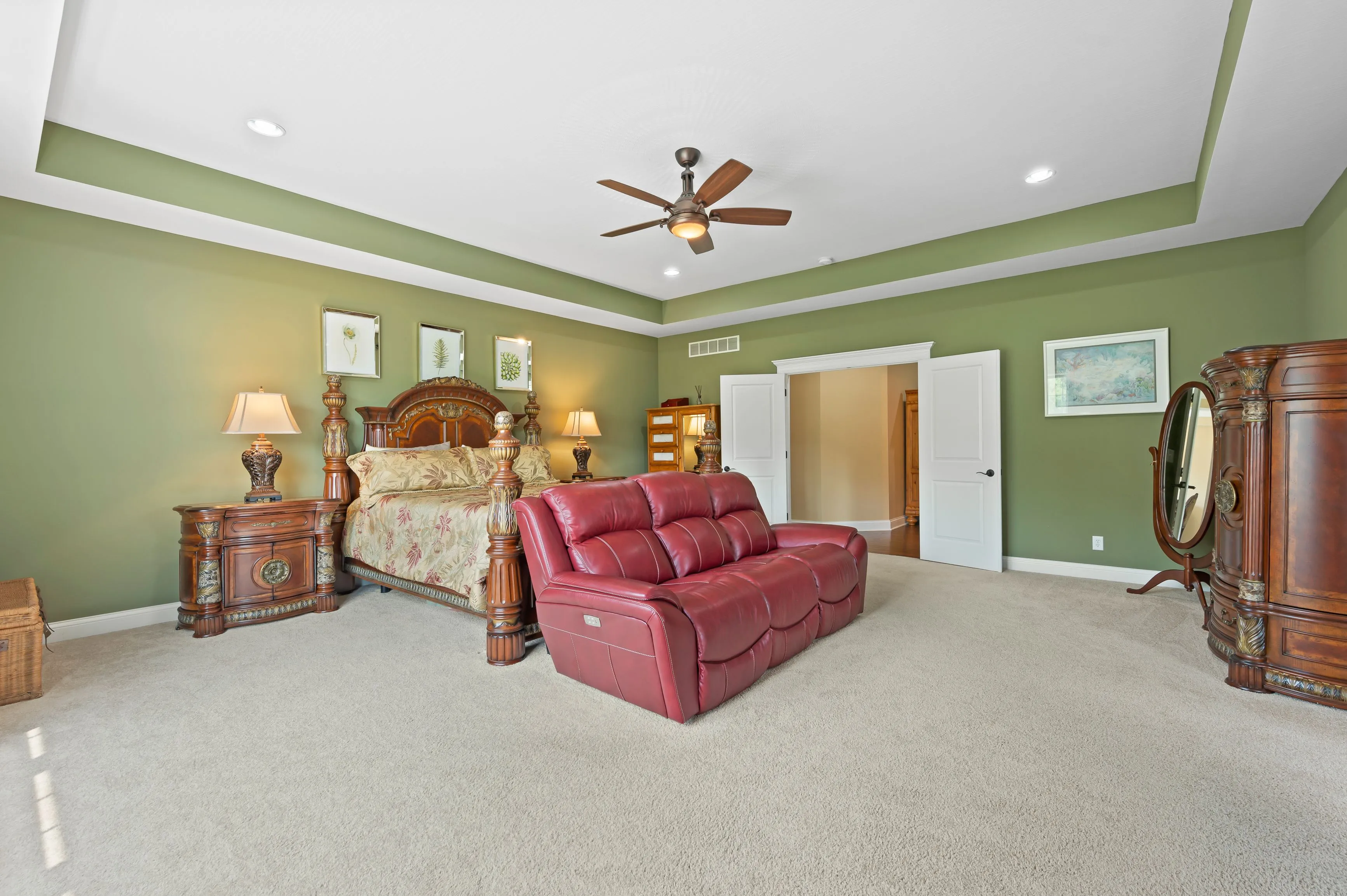 Spacious bedroom with green walls, a large bed with an ornate headboard, red leather sofa, wooden furniture, a ceiling fan, and cream carpet flooring.