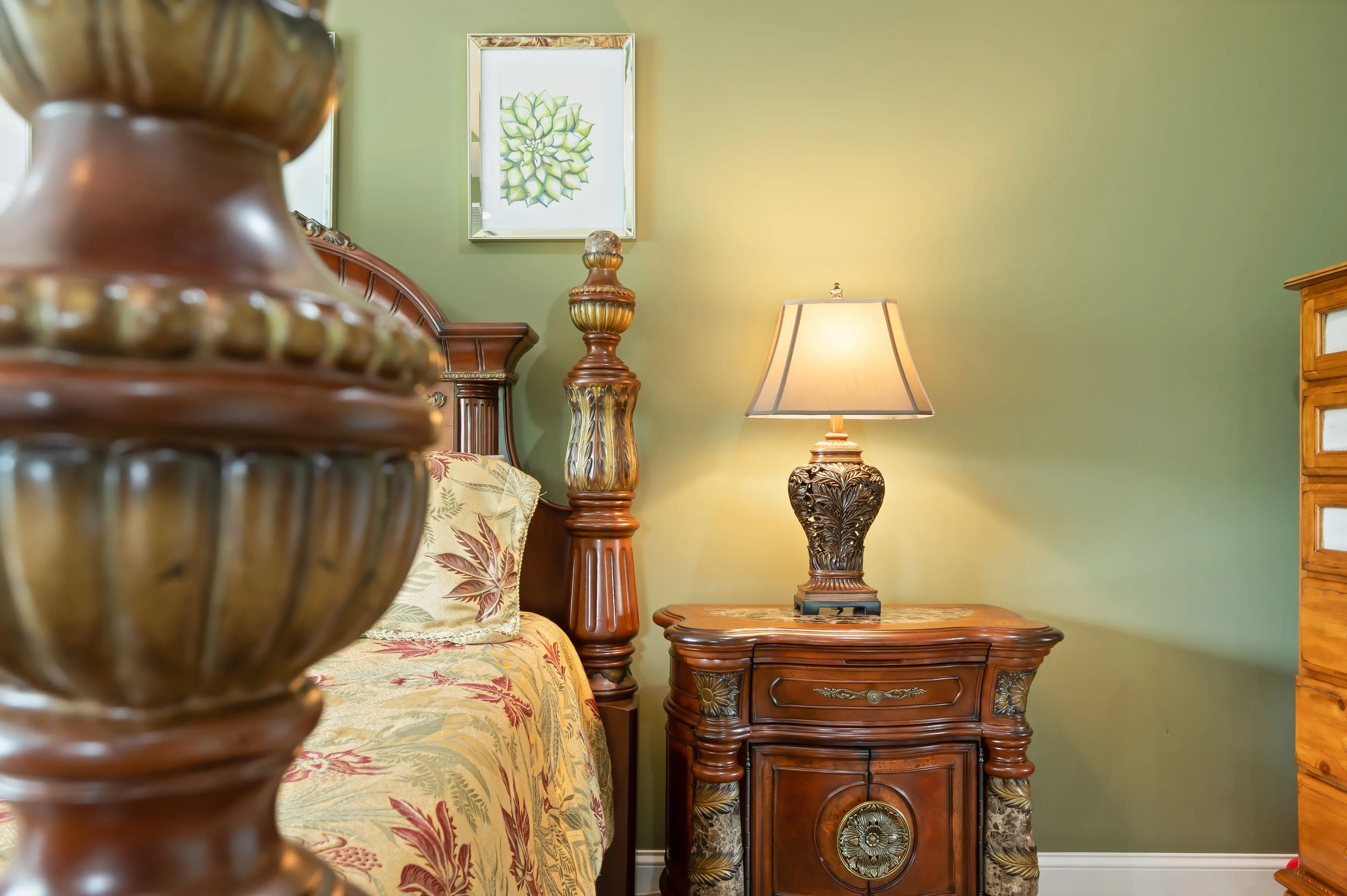 Elegant bedroom interior with carved wooden furniture, ornate table lamp, and artwork on green wall.