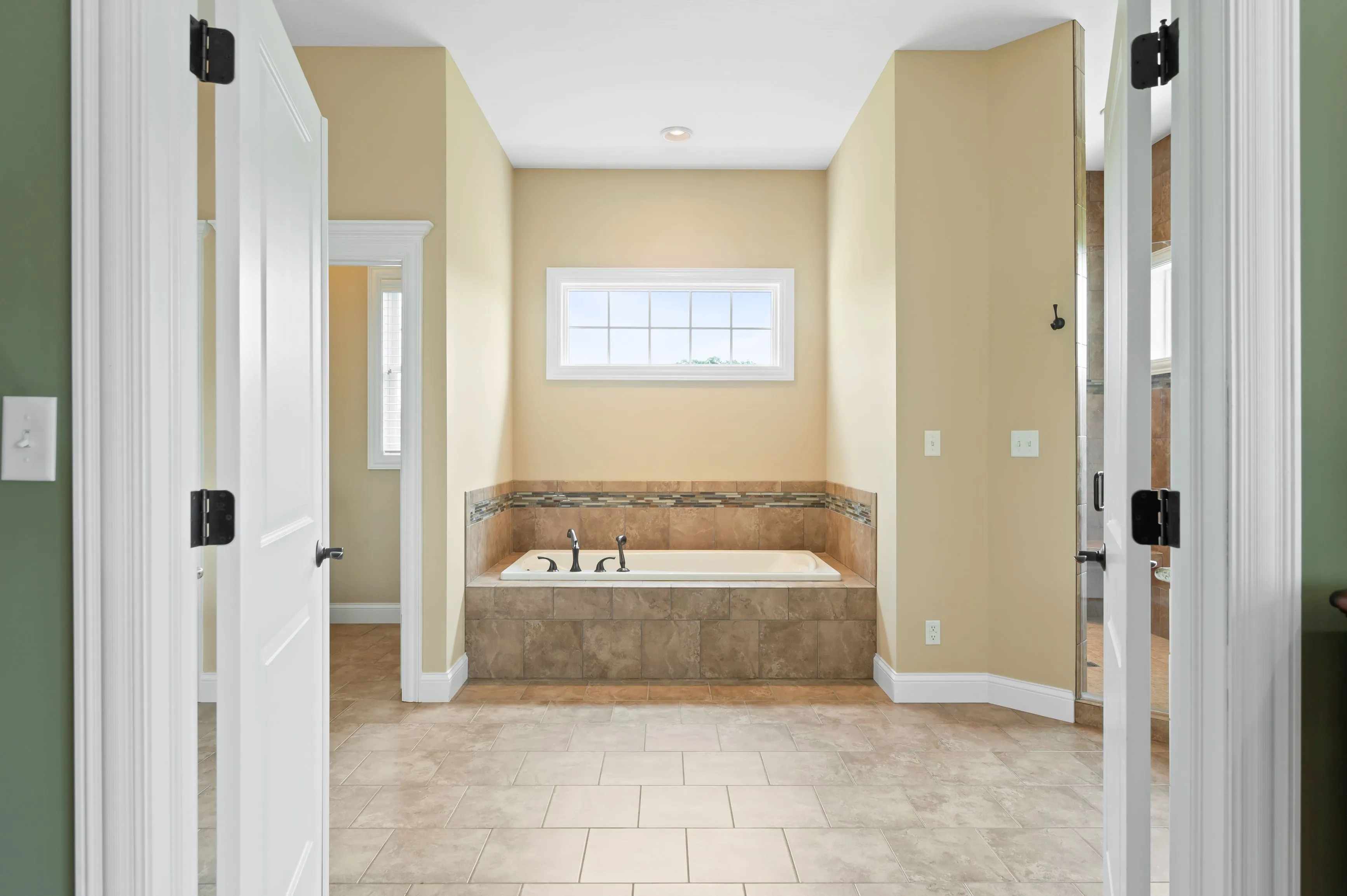 A spacious bathroom with beige walls, tiled flooring, a large built-in bathtub with tiled surround, seen through an open door.