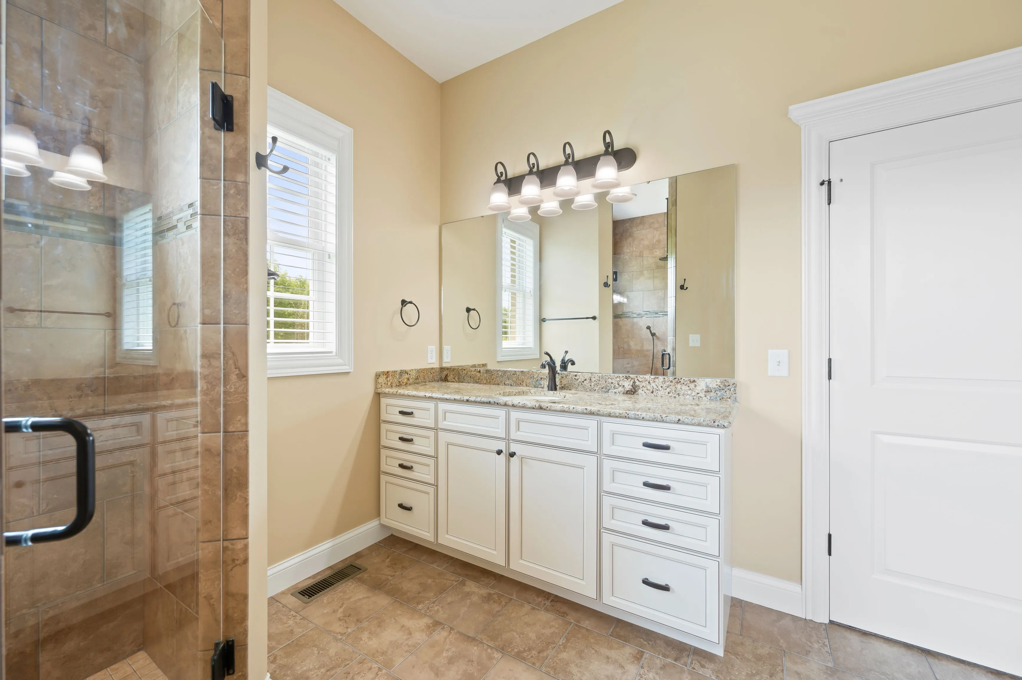 Modern bathroom interior with dual sinks, granite countertops, large mirror, and a glass-enclosed shower.