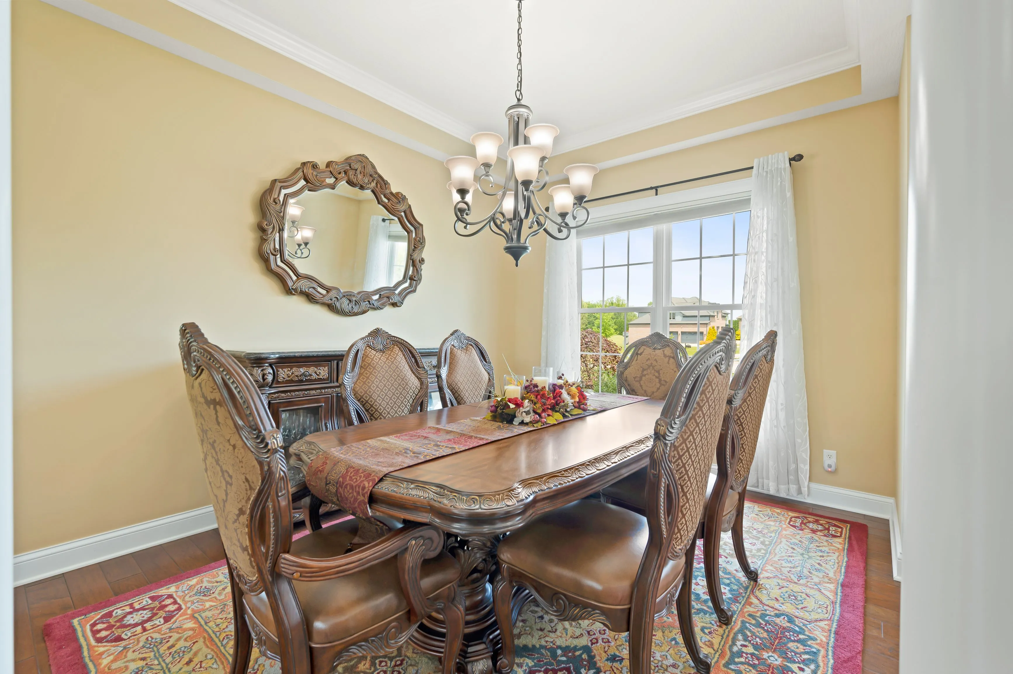 Elegant dining room with a polished wooden table, ornate chairs, and a decorative mirror on the wall, bathed in natural light from a window with sheer curtains.