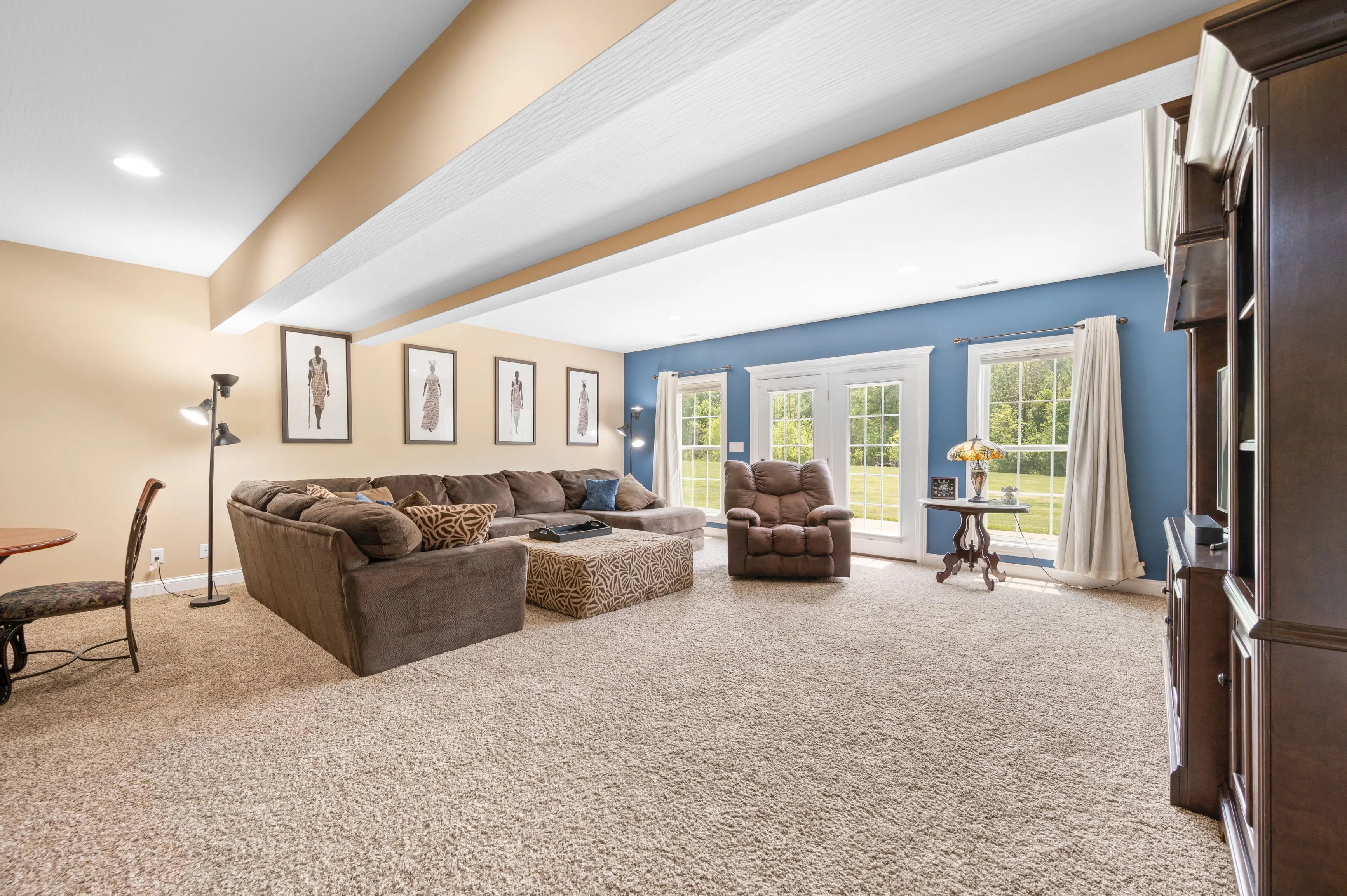 Spacious living room with beige carpeting, large brown sectional sofa, recliner, and framed artwork on the walls, accented by blue-painted walls and white trim with ample natural light from windows.