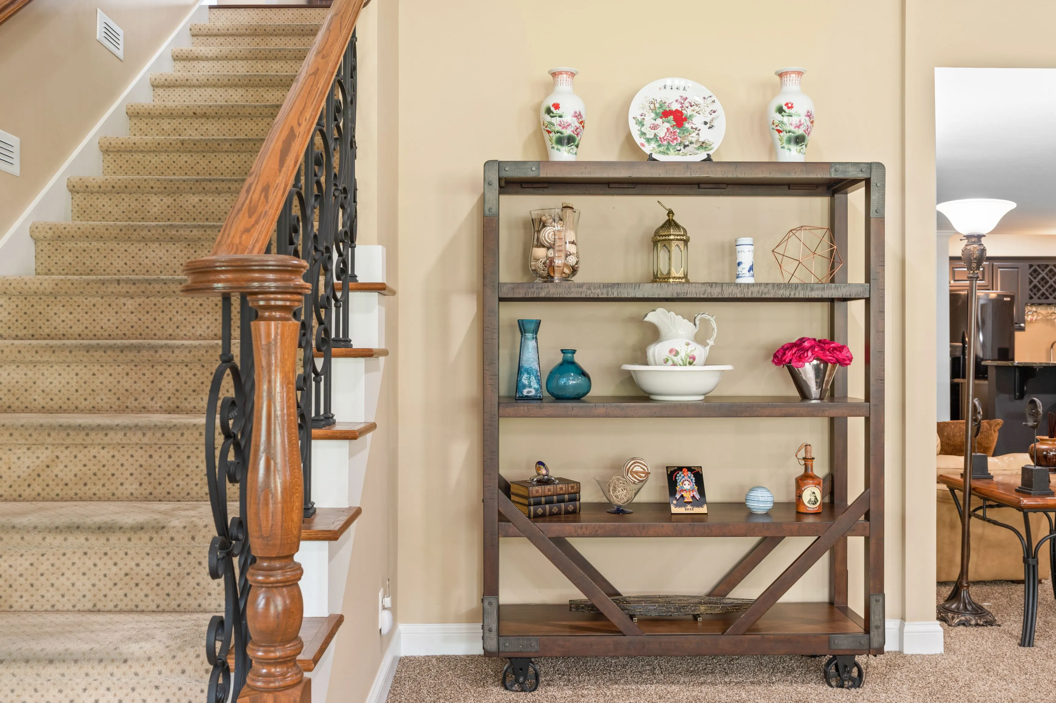 Elegant home interior featuring a wooden staircase with ornate iron balusters and a wooden shelf cart displaying various decorative items such as vases, plates, and figurines.