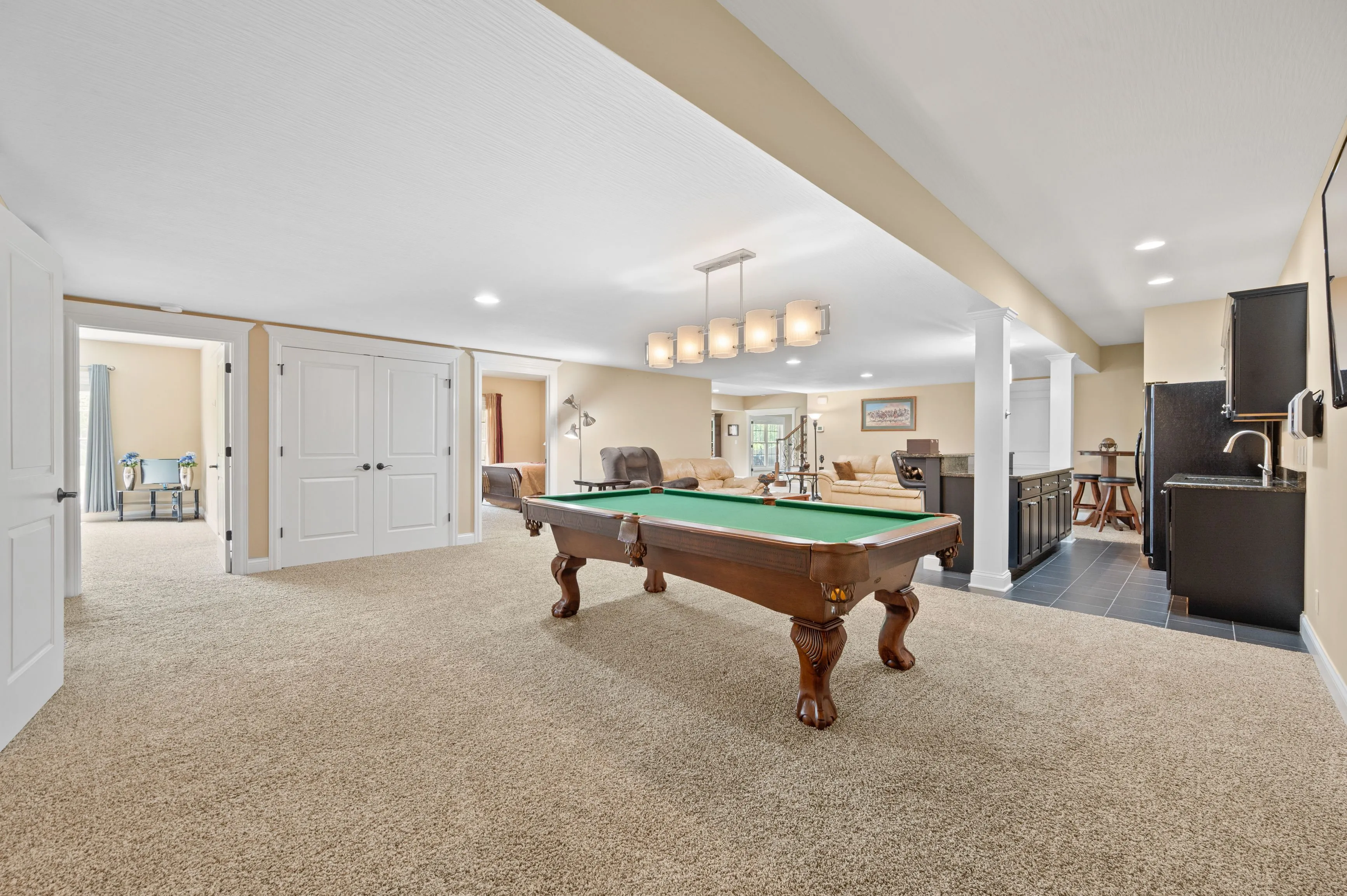 Spacious basement recreation room with a pool table, beige carpet, white ceiling, and beige walls, including a seating area and a minibar.