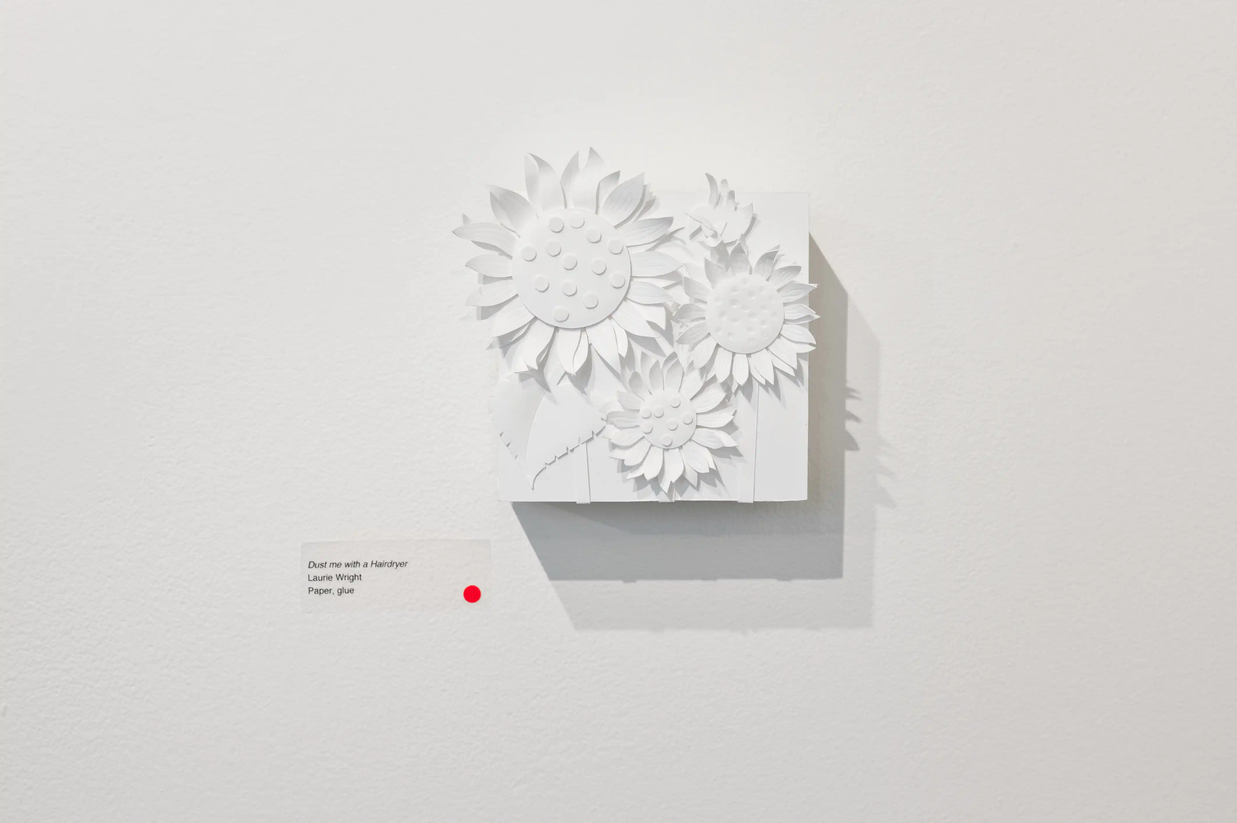 White three-dimensional floral wall art installation with a description label and a red security sensor on a white wall.