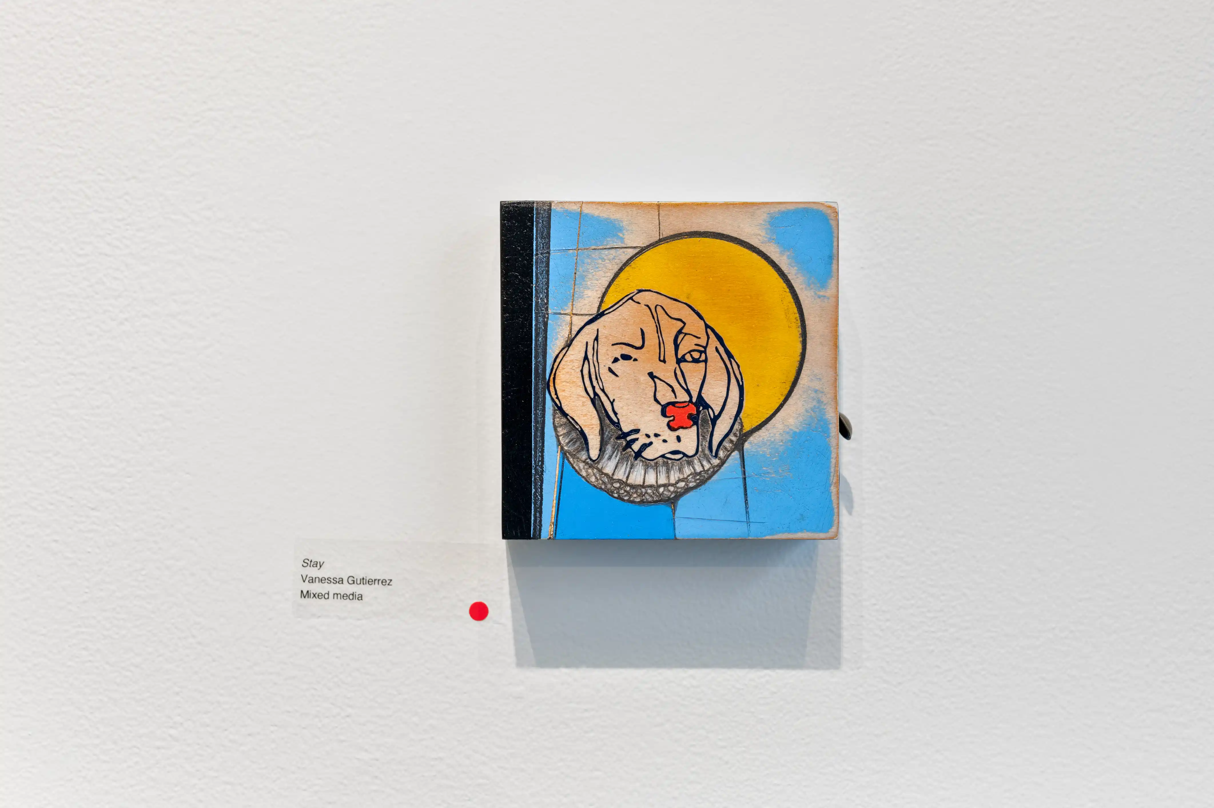 Artwork on display featuring a stylized face with a yellow halo on a blue background, exhibited on a white gallery wall with an information label on the left and a red dot on the right.