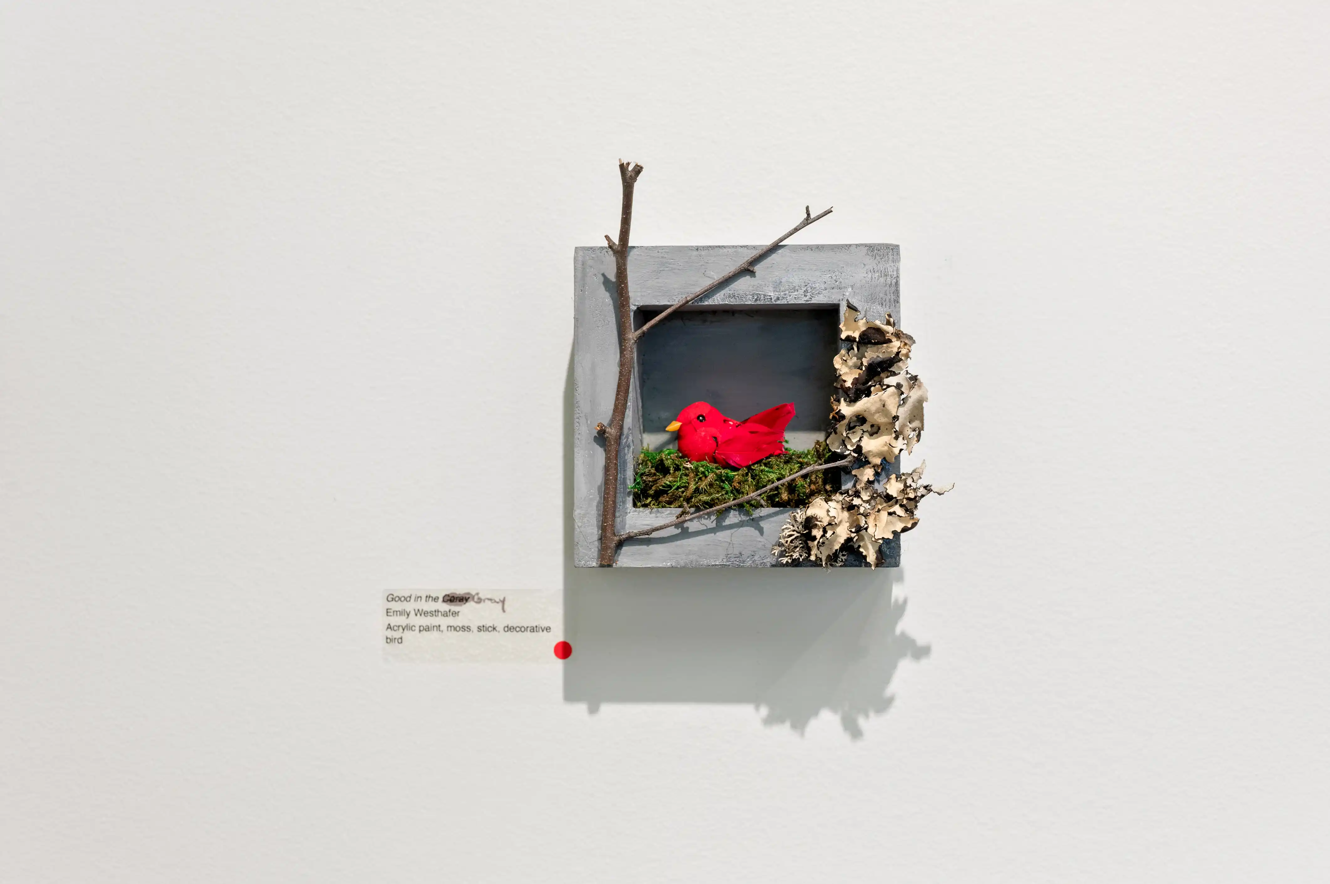 Art installation of a gray cube on a white wall with natural elements and a red fabric heart inside.