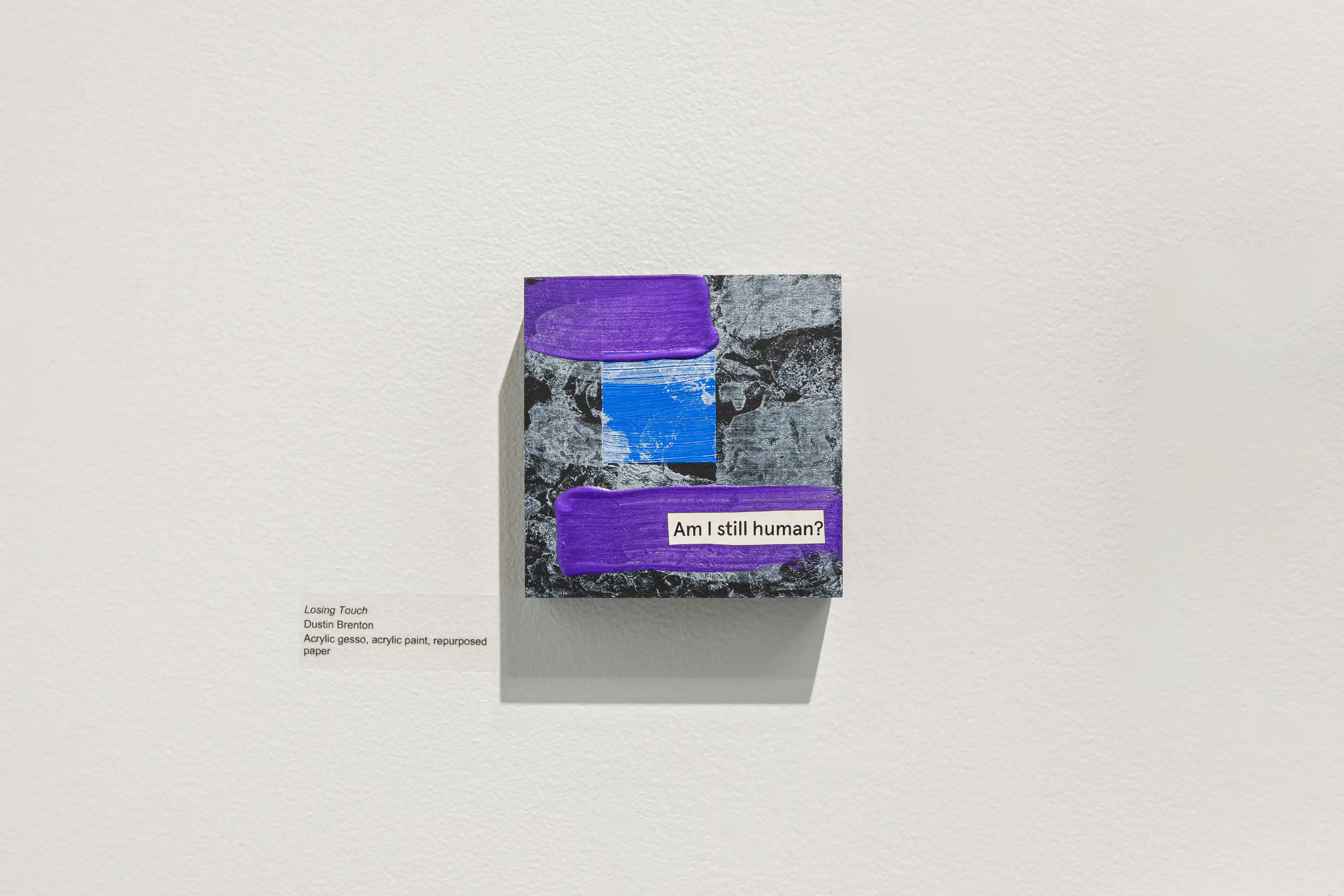 Art book displayed on a white wall with purple and gray abstract cover design, title partially obscured, mounted with descriptive text plaque beside it.