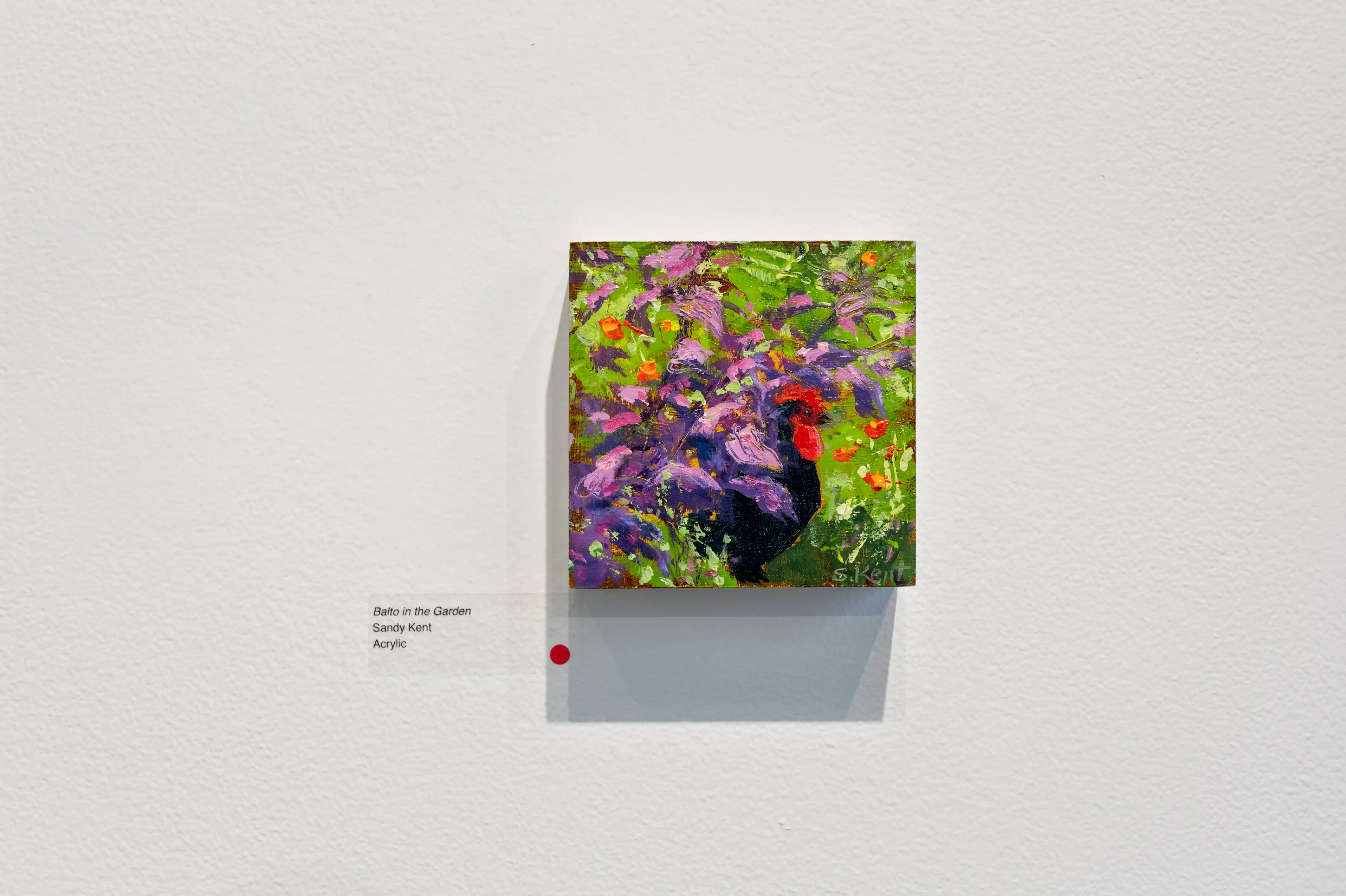 Painting of a colorful rooster among flowers on display in an art gallery with a gallery label on the wall.