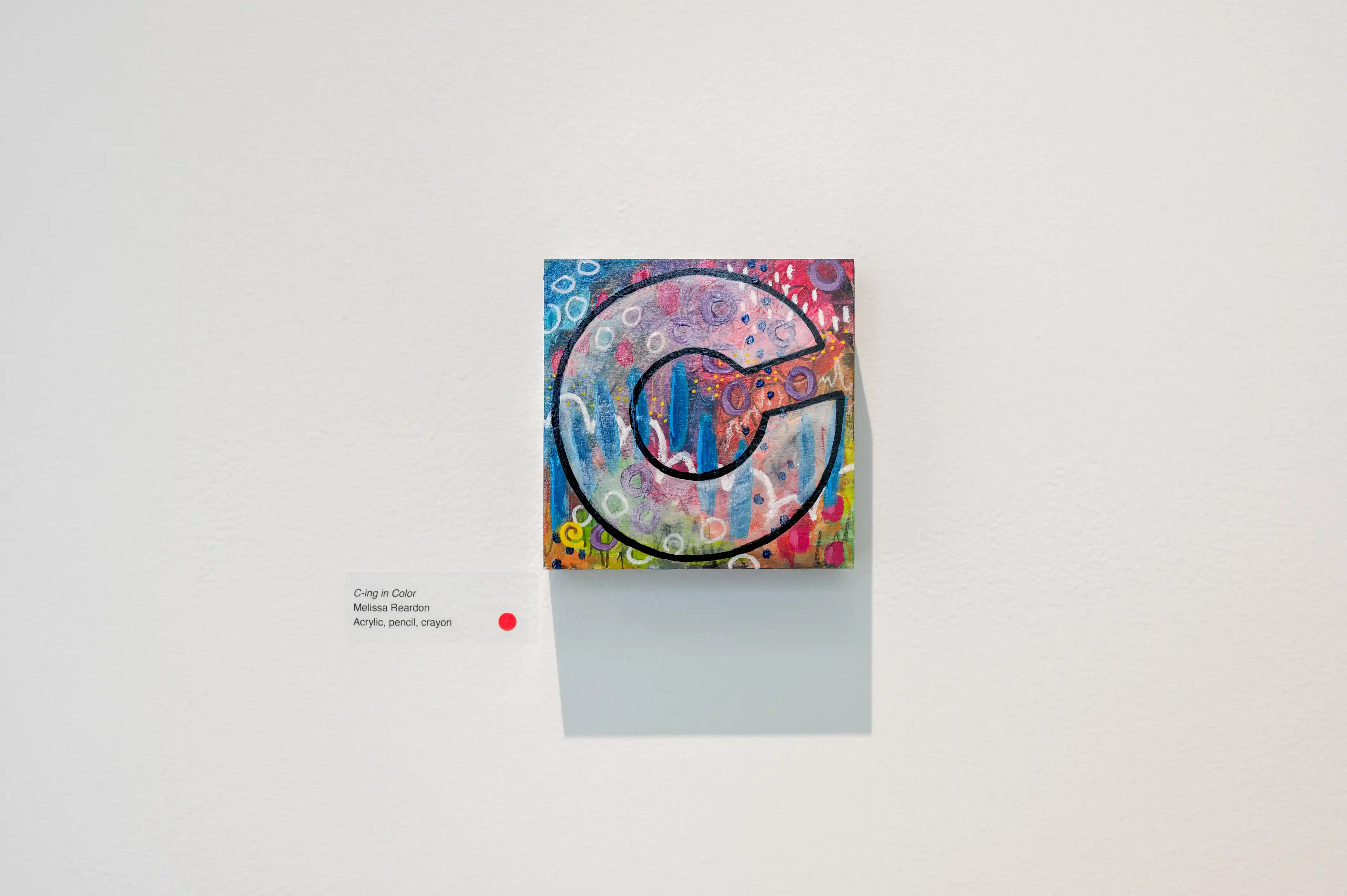 Colorful painting of a stylized "G" on a canvas displayed on a white gallery wall with descriptive text label to the side.