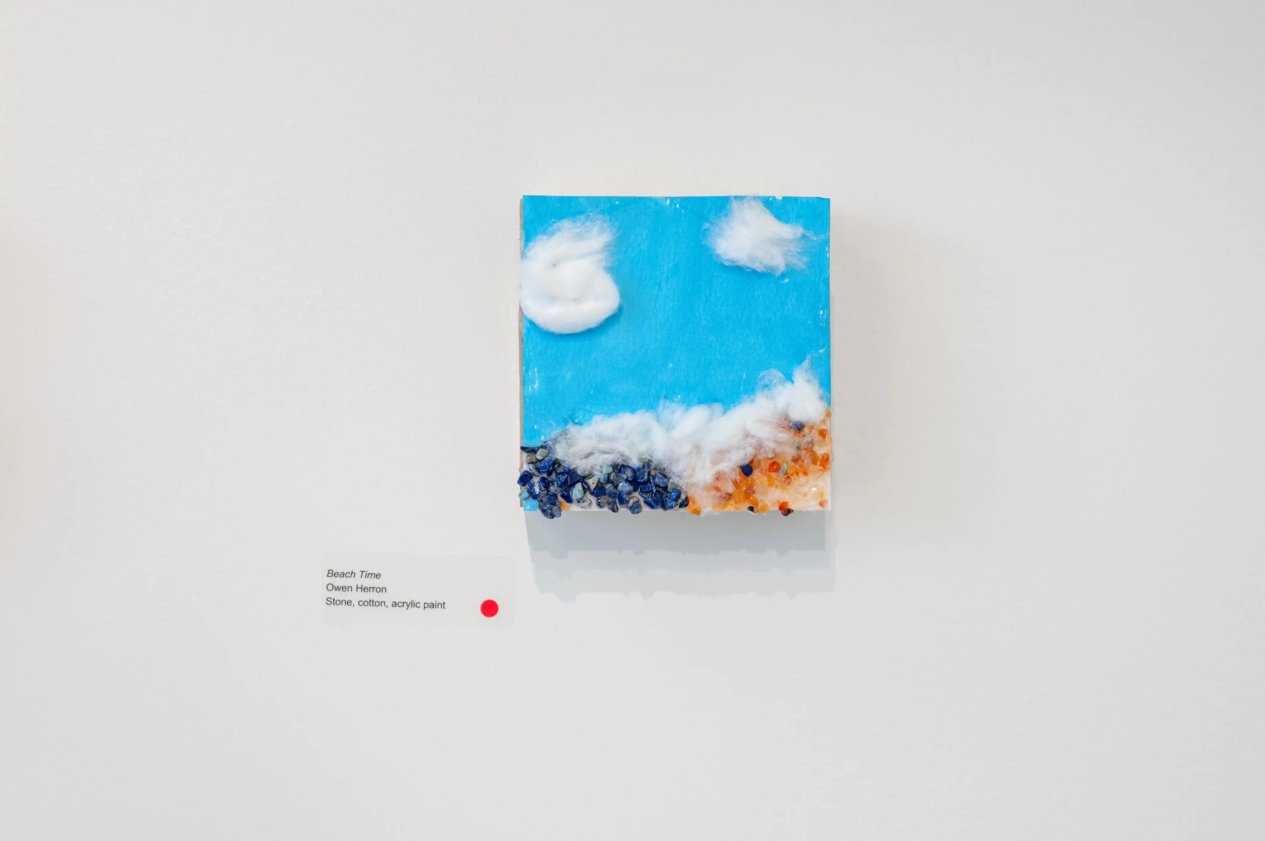 Small abstract painting with a blue sky and white clouds on upper part and rough orange texture on the lower part, displayed on a white wall with a red dot and title card indicating the piece is sold.