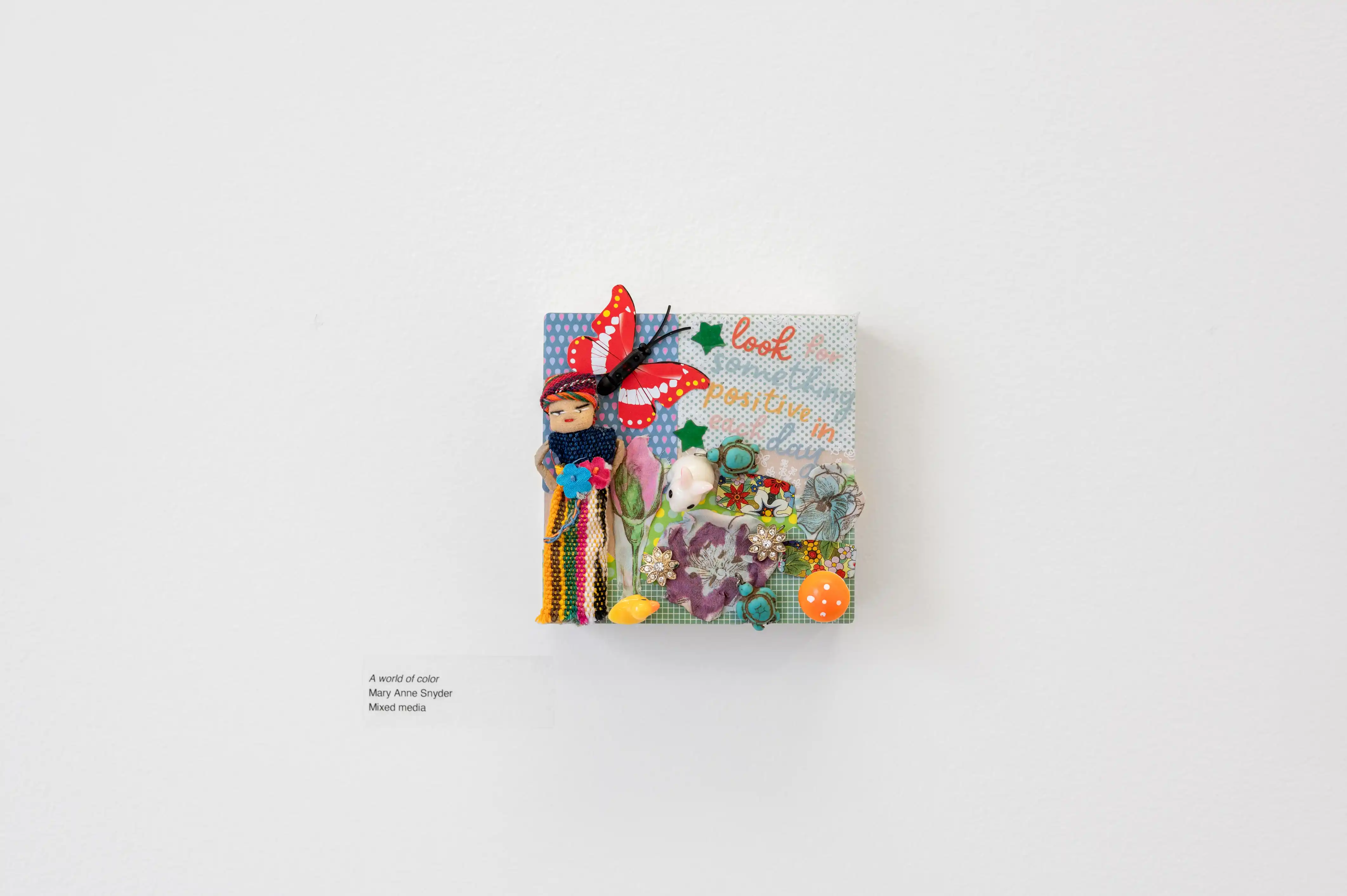 Art installation on a white wall featuring a colorful collage of assorted objects, paper cutouts, and ribbons with handwritten text at the bottom.