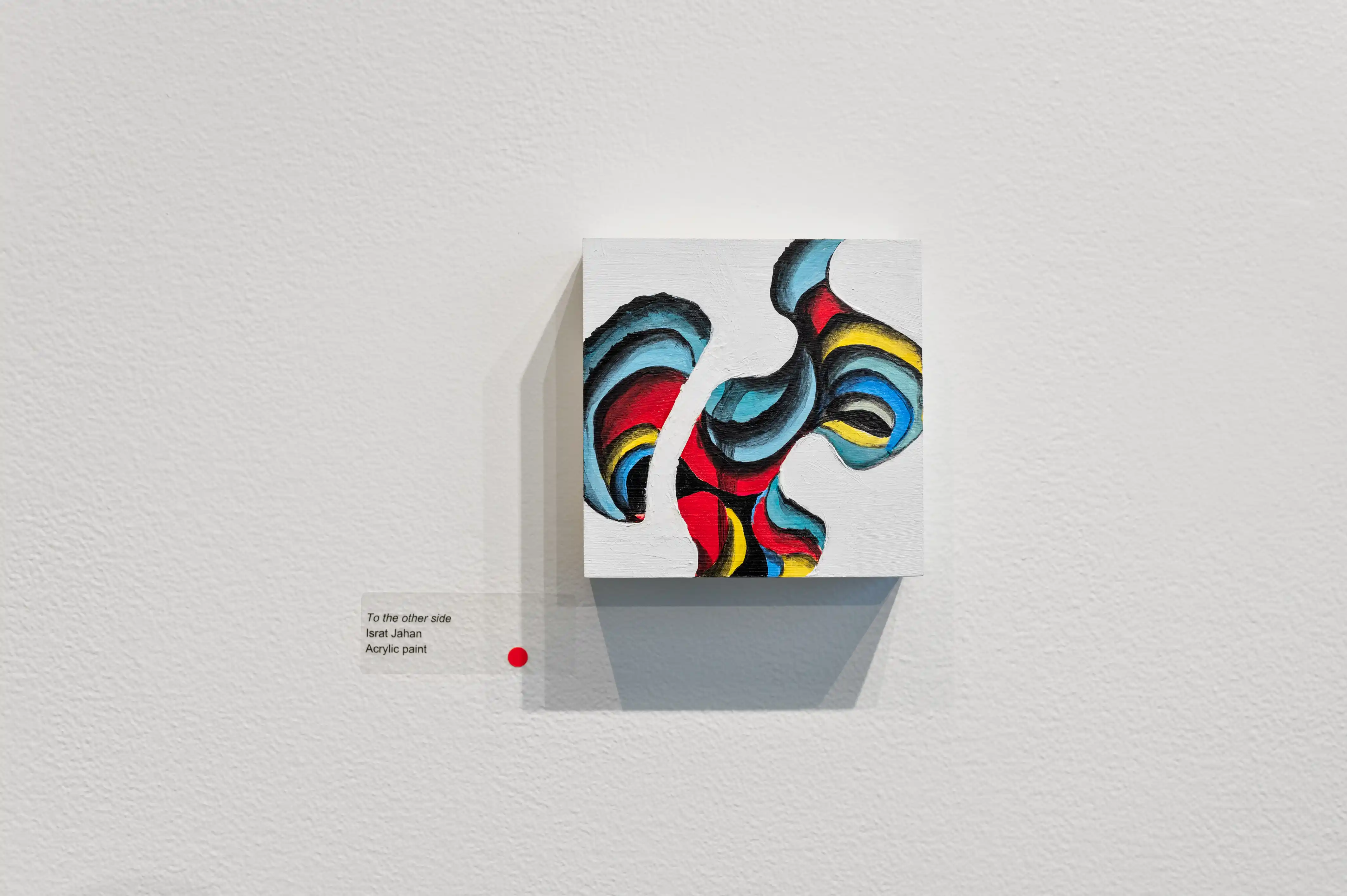 Abstract multicolored sculpture displayed on a white pedestal against a white wall with exhibition details on a small label to the side.