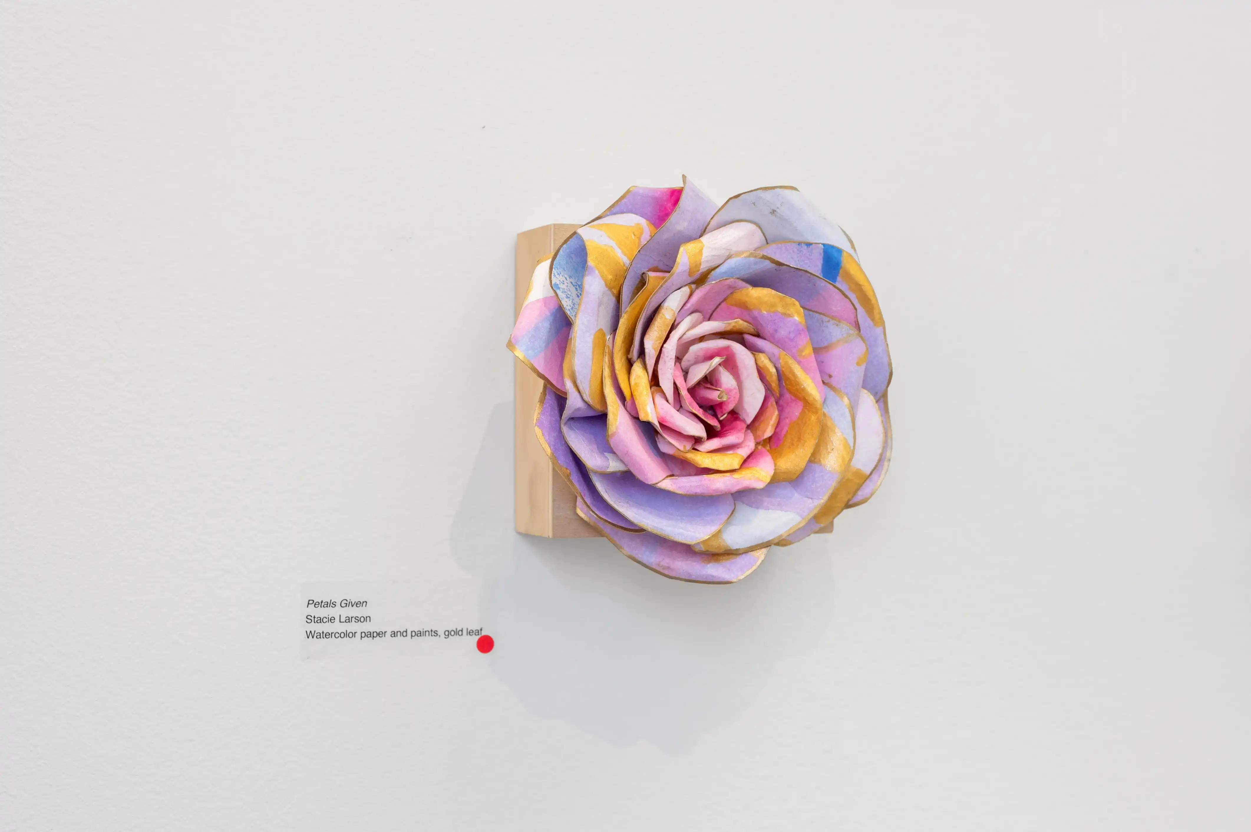 Decorative paper flower with pastel colors mounted on a white wall, with an artist's signature at the bottom left.