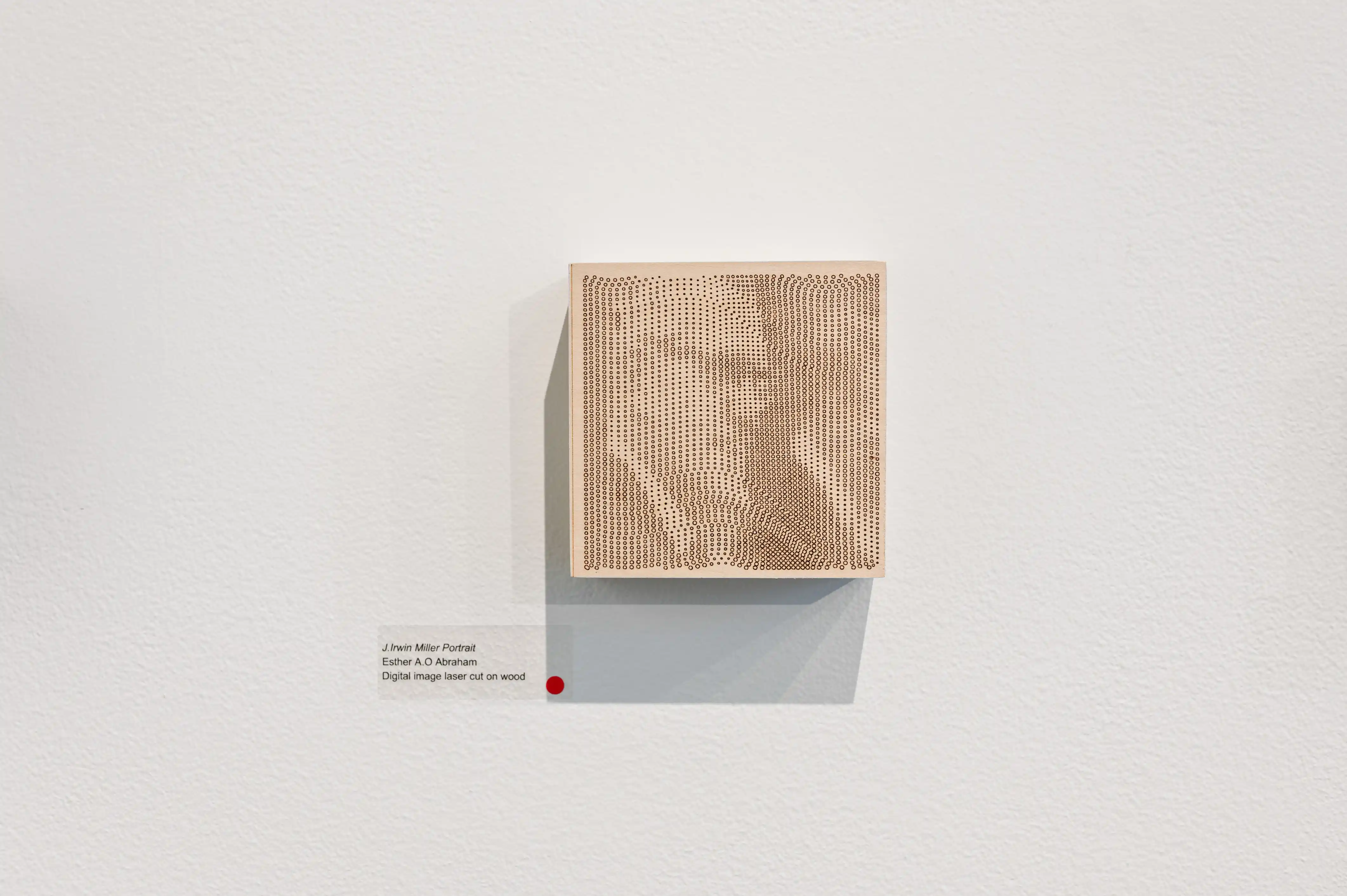 A minimalist art installation featuring a single, textured beige cube mounted on a white wall with a small red detail to the bottom left corner and a descriptive label at the bottom right.