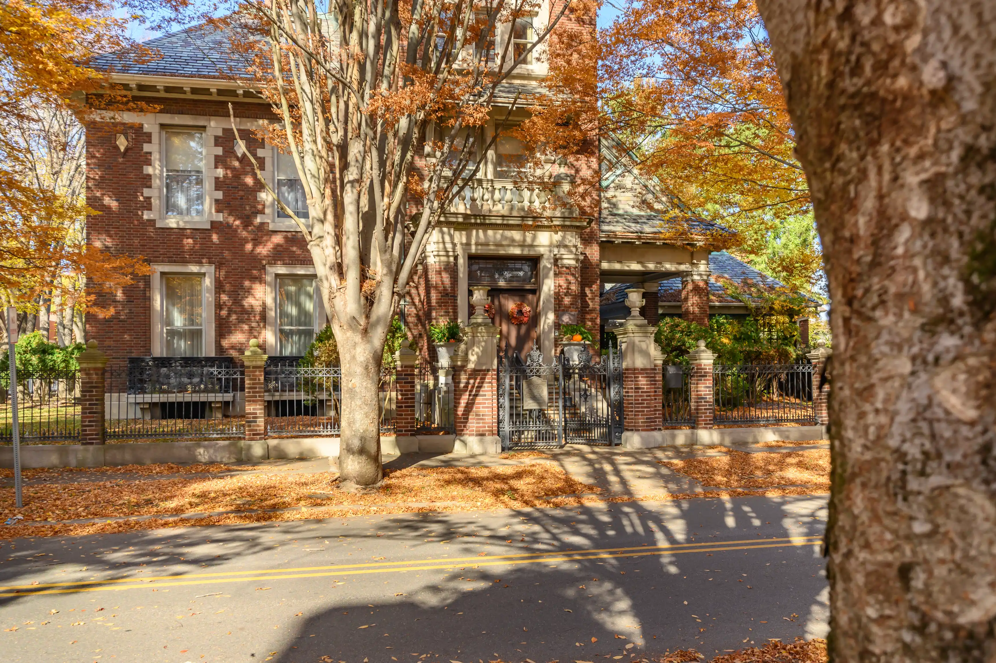 Brick house with autumn foliage, iron fence, and fallen leaves on a sunny day.