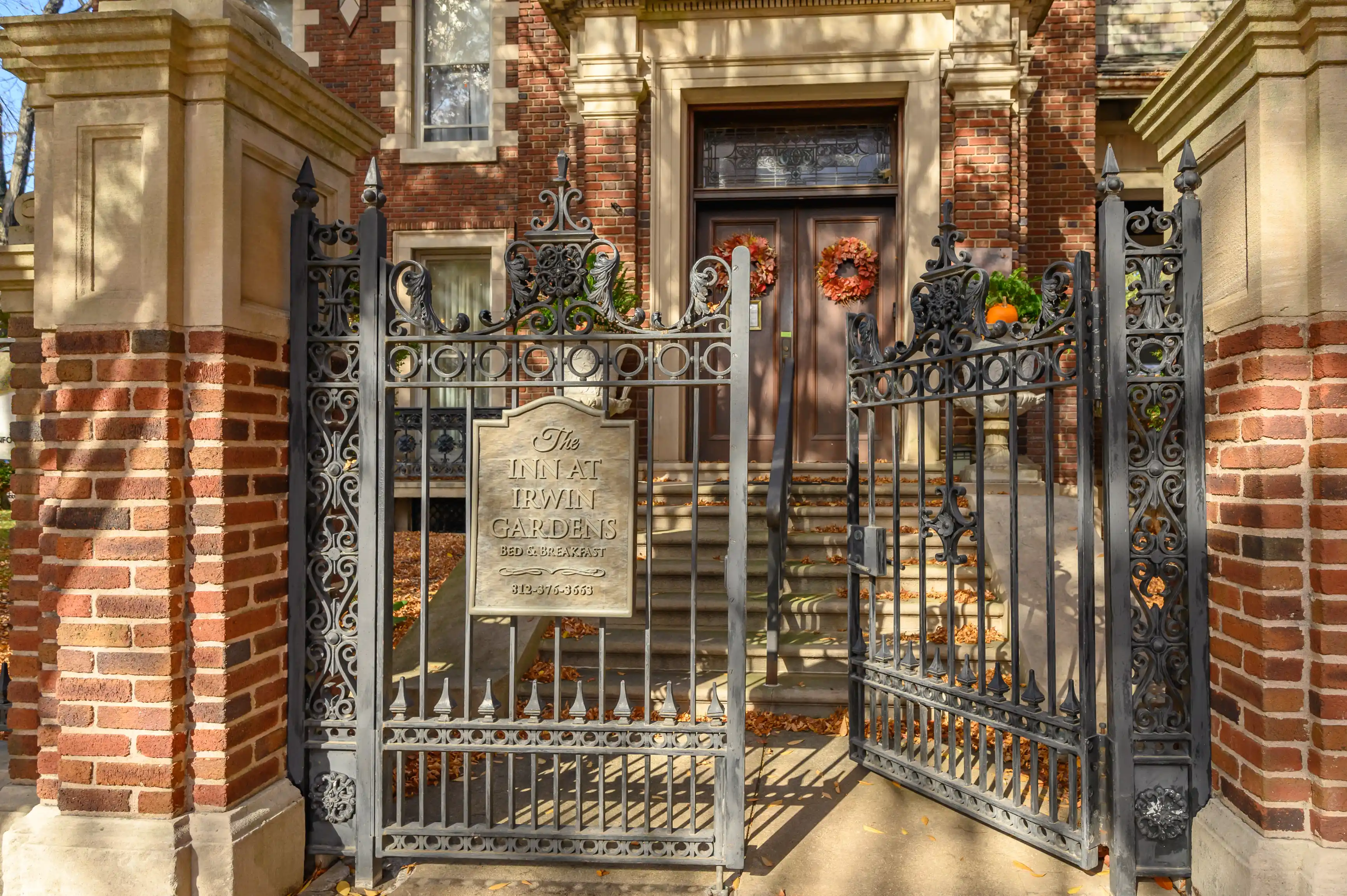 Ornate wrought iron gate open inwards towards the entrance of "The Inn at Irwin Gardens Bed & Breakfast" with decorative wreaths on doors and a signboard with contact information on a sunny day.
