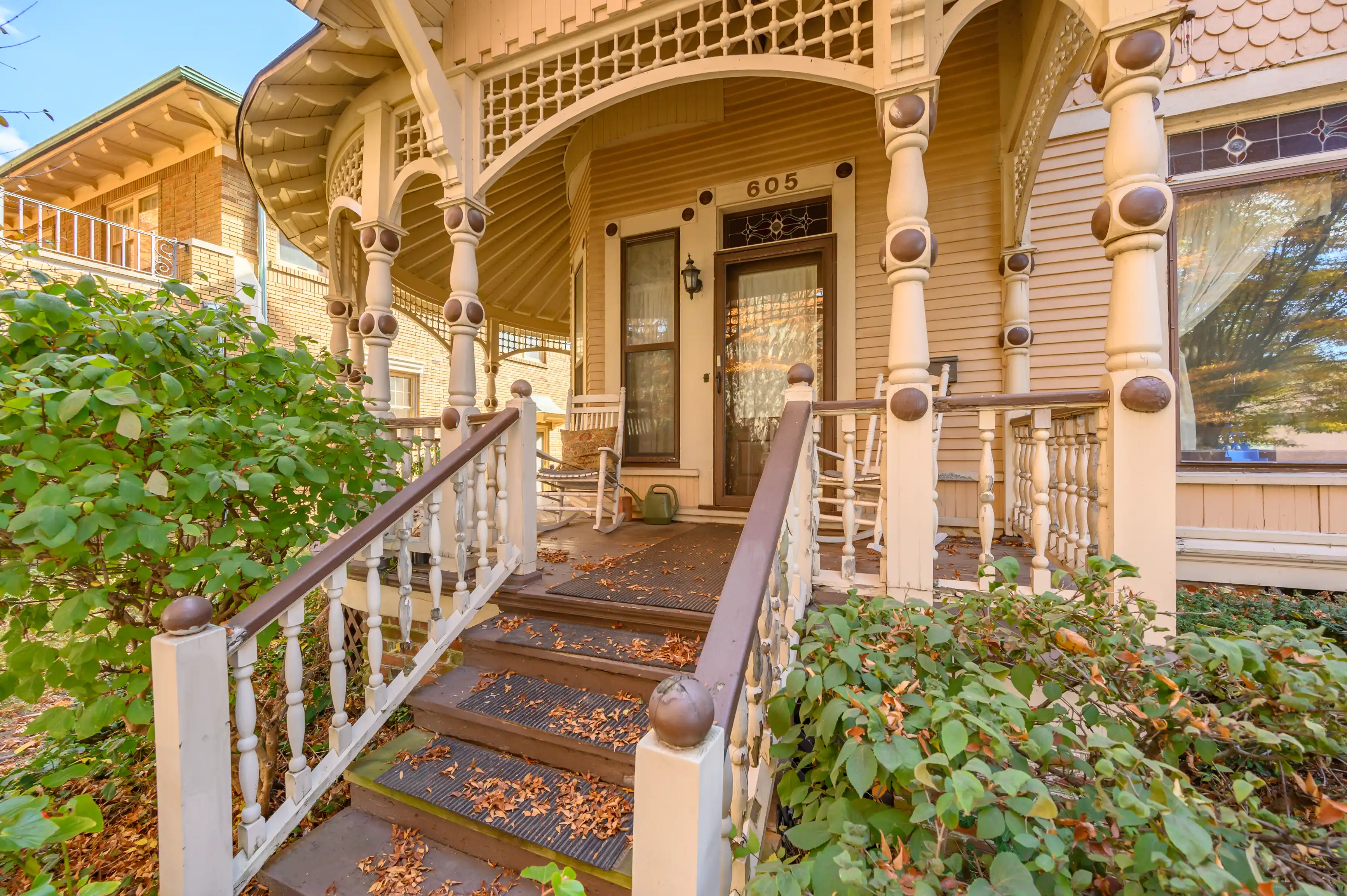 Charming Victorian style home entrance with an inviting porch featuring ornate wooden columns, decorative railings, and steps with scattered autumn leaves.
