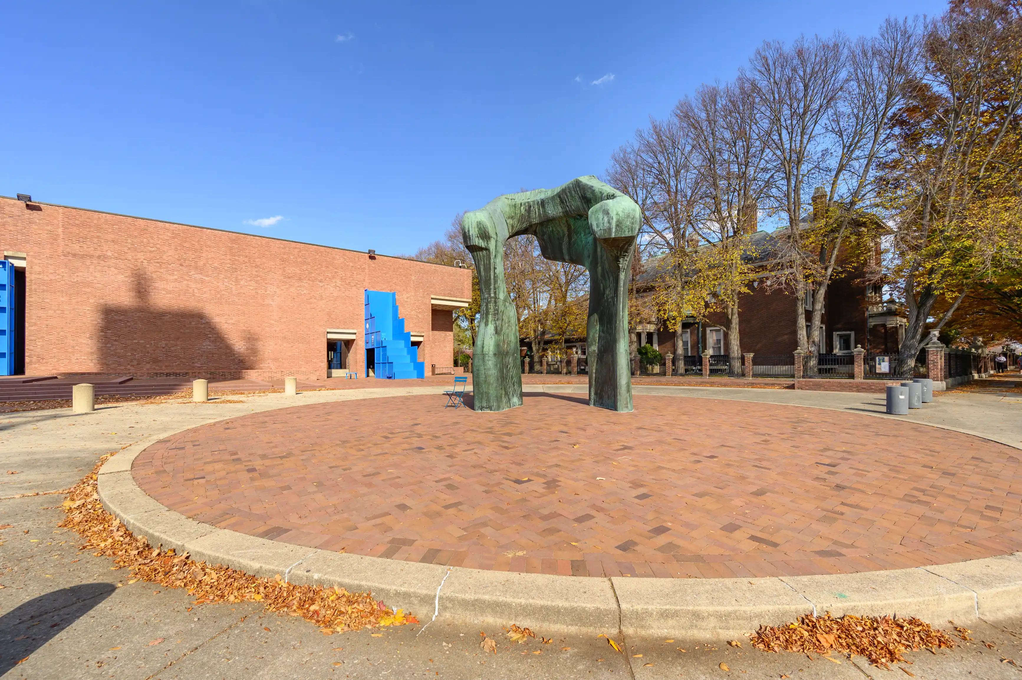 Large bronze sculpture in a public plaza with brick pavement, surrounded by trees and a building with bright blue elements in the background.