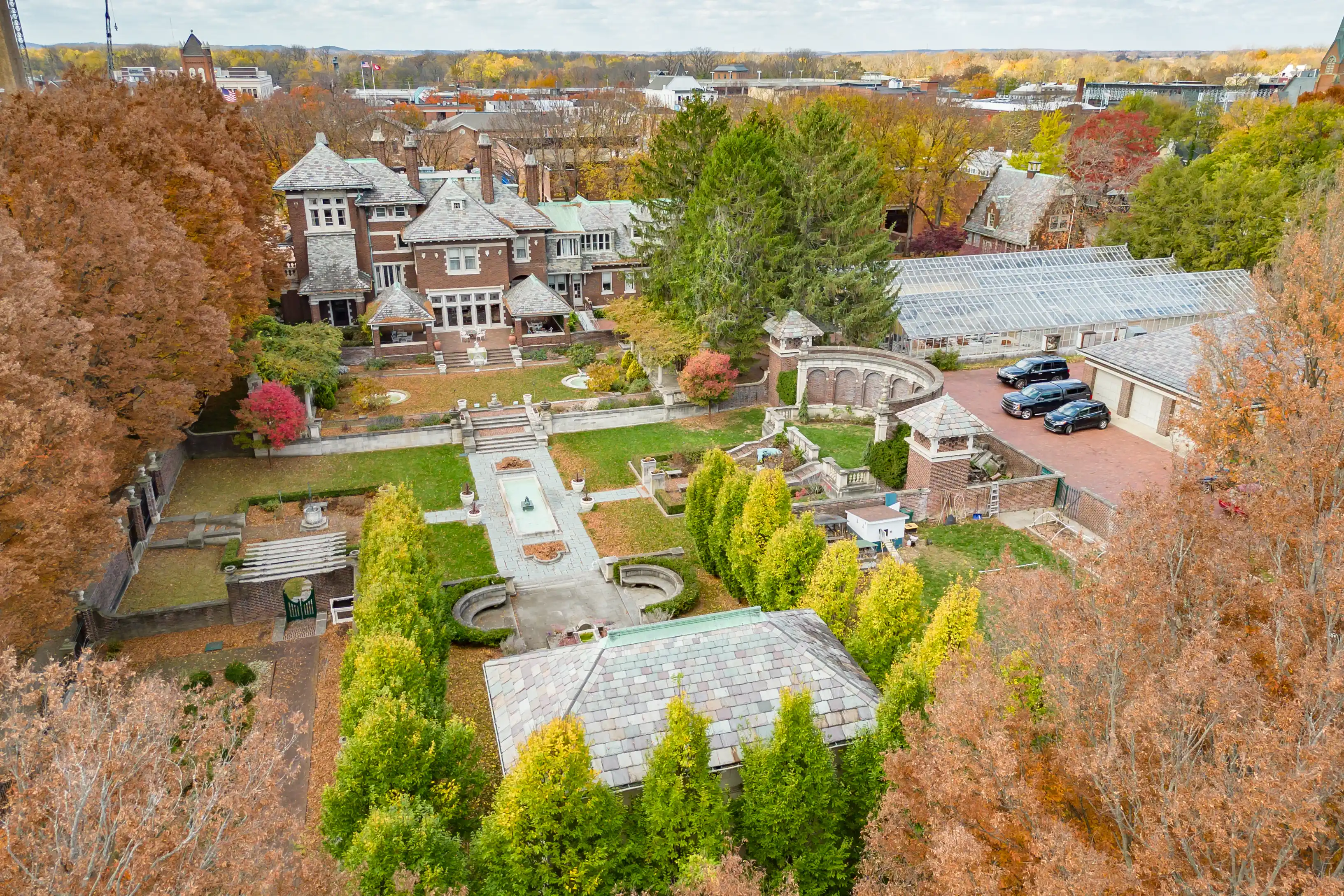 Aerial view of a stately historic mansion with formal gardens and autumn-colored trees, with a greenhouse and a town in the background.