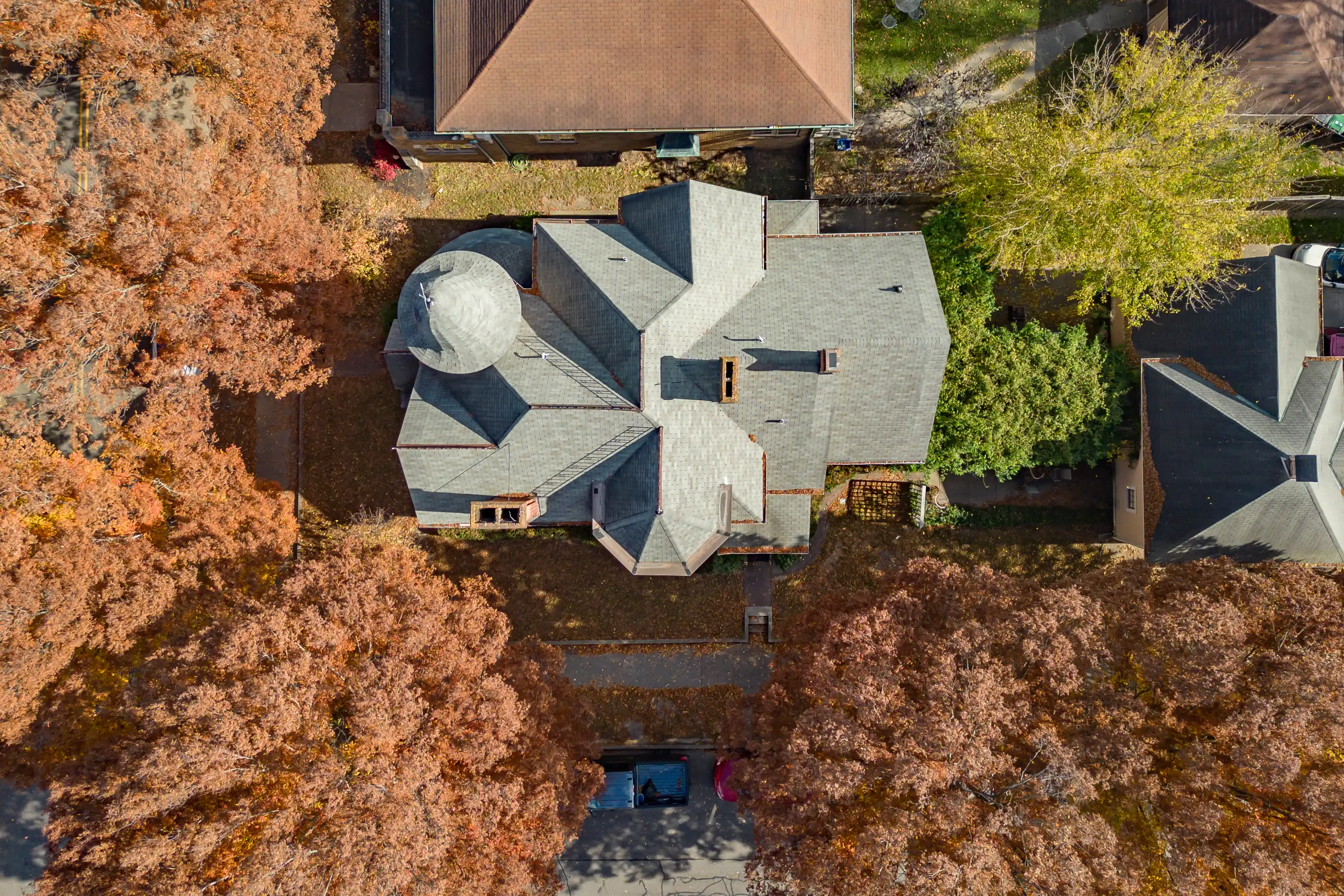 Aerial view of a residential house with a complex gray roof and surrounding autumn-colored trees.