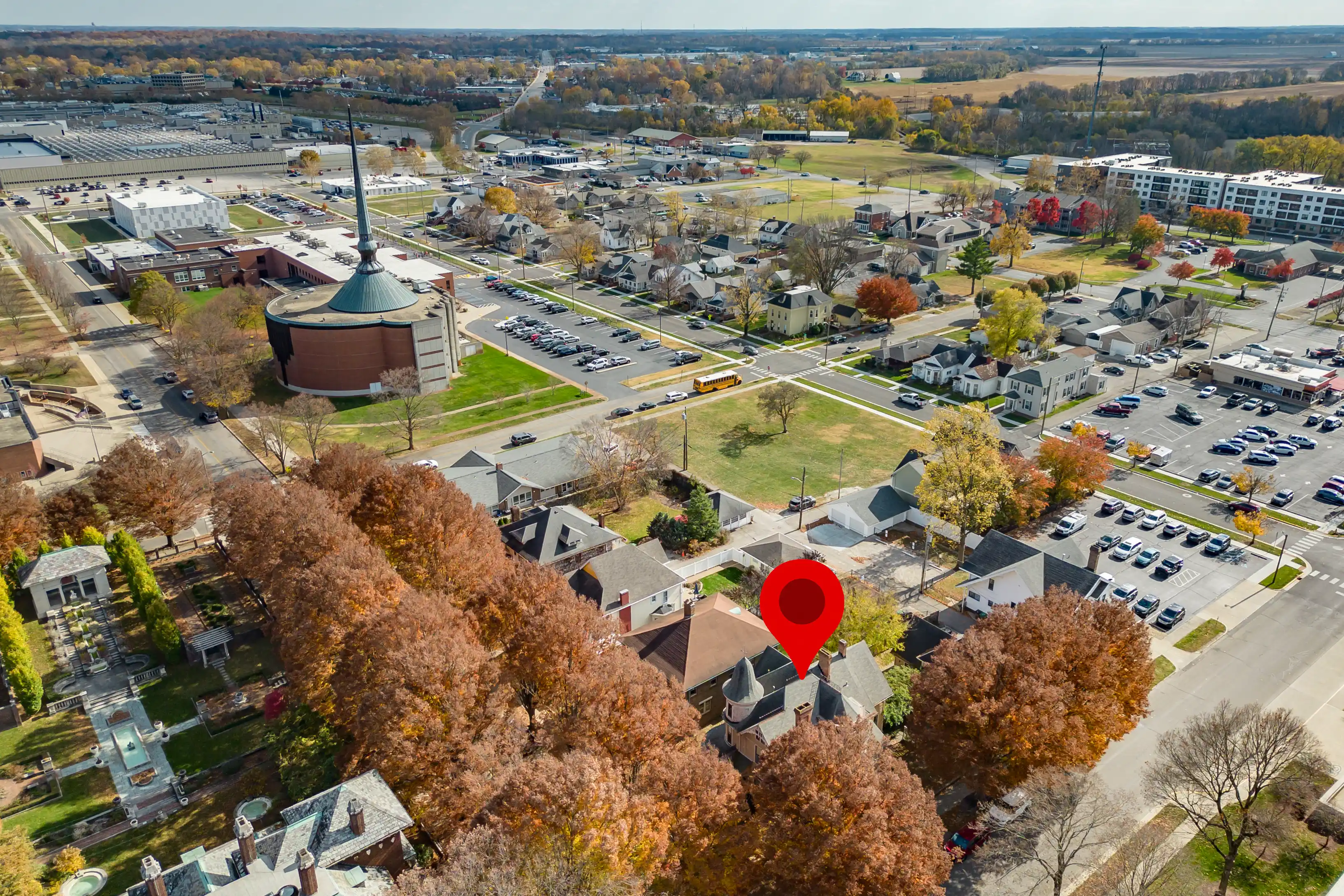 Aerial view of a suburban area with houses, trees showing autumn foliage, a round building with a spire, a red location marker over one of the houses, and a large parking area in the distance.
