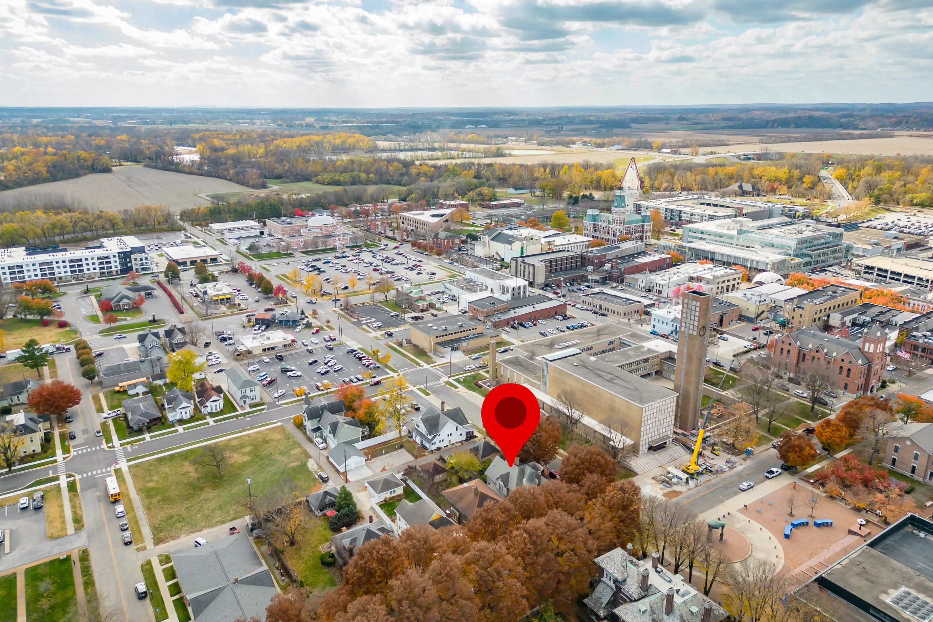 Aerial view of a suburban town with mixed residential and commercial buildings, featuring a red location marker pin over a specific building, amidst autumn-colored trees and overcast skies.