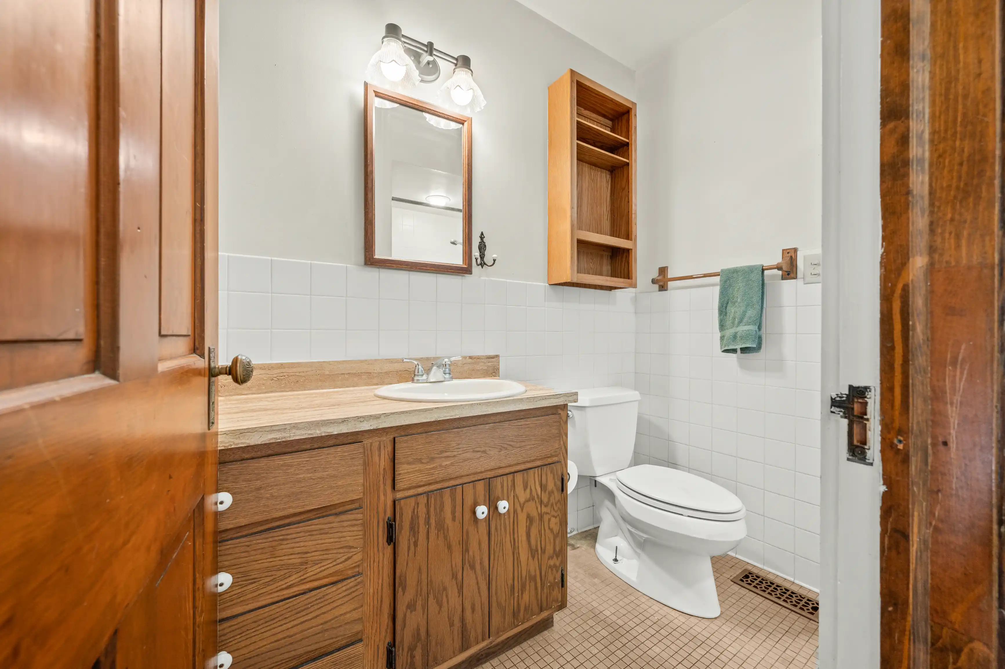 Interior of a small bathroom with wooden vanity, white sink, mirror with overhead light, toilet, white tiling, and wooden open wall cabinet. A green towel hangs on a rack by the door, which is ajar.