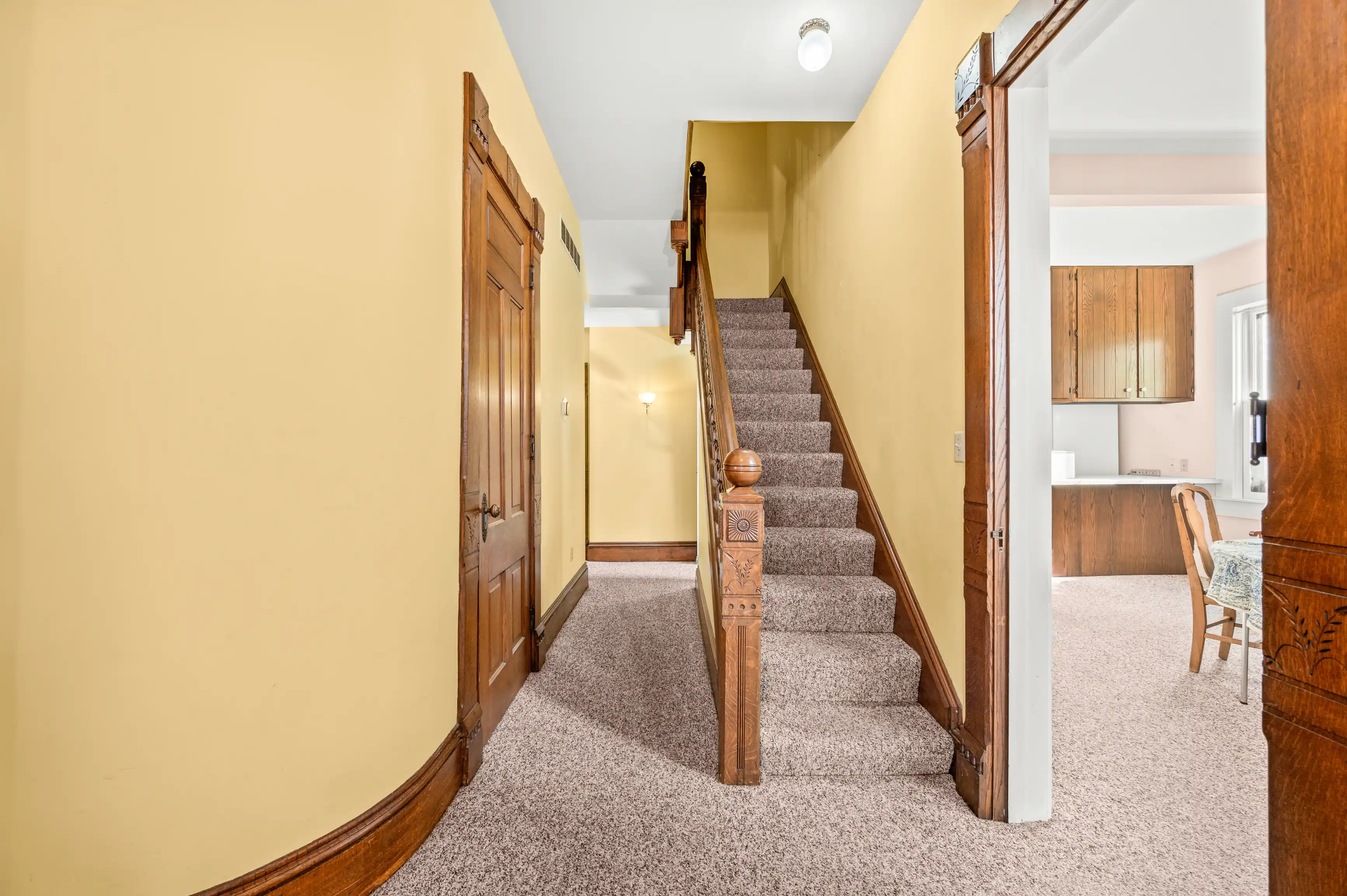 Interior view of a home showing a carpeted staircase with wooden banister, a hallway leading to other rooms, and an open door to a kitchen with wooden cabinets.