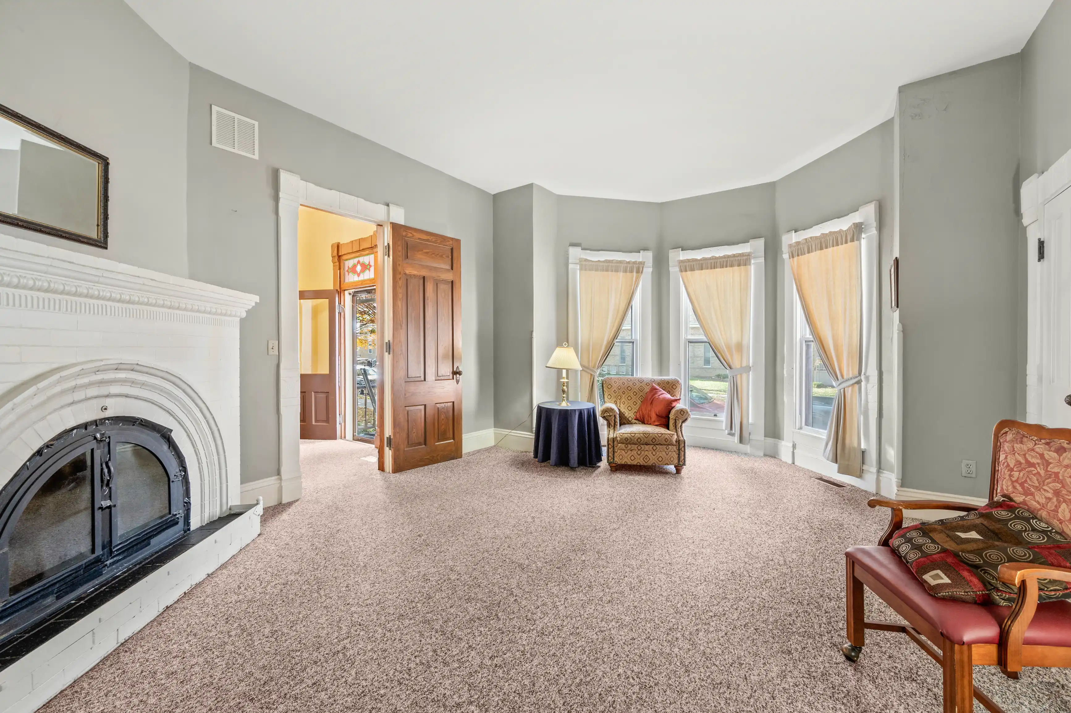 Spacious living room with plush carpet, a white fireplace, bay windows with curtains, classic wooden furniture, and a view into a hallway with a stained glass door.