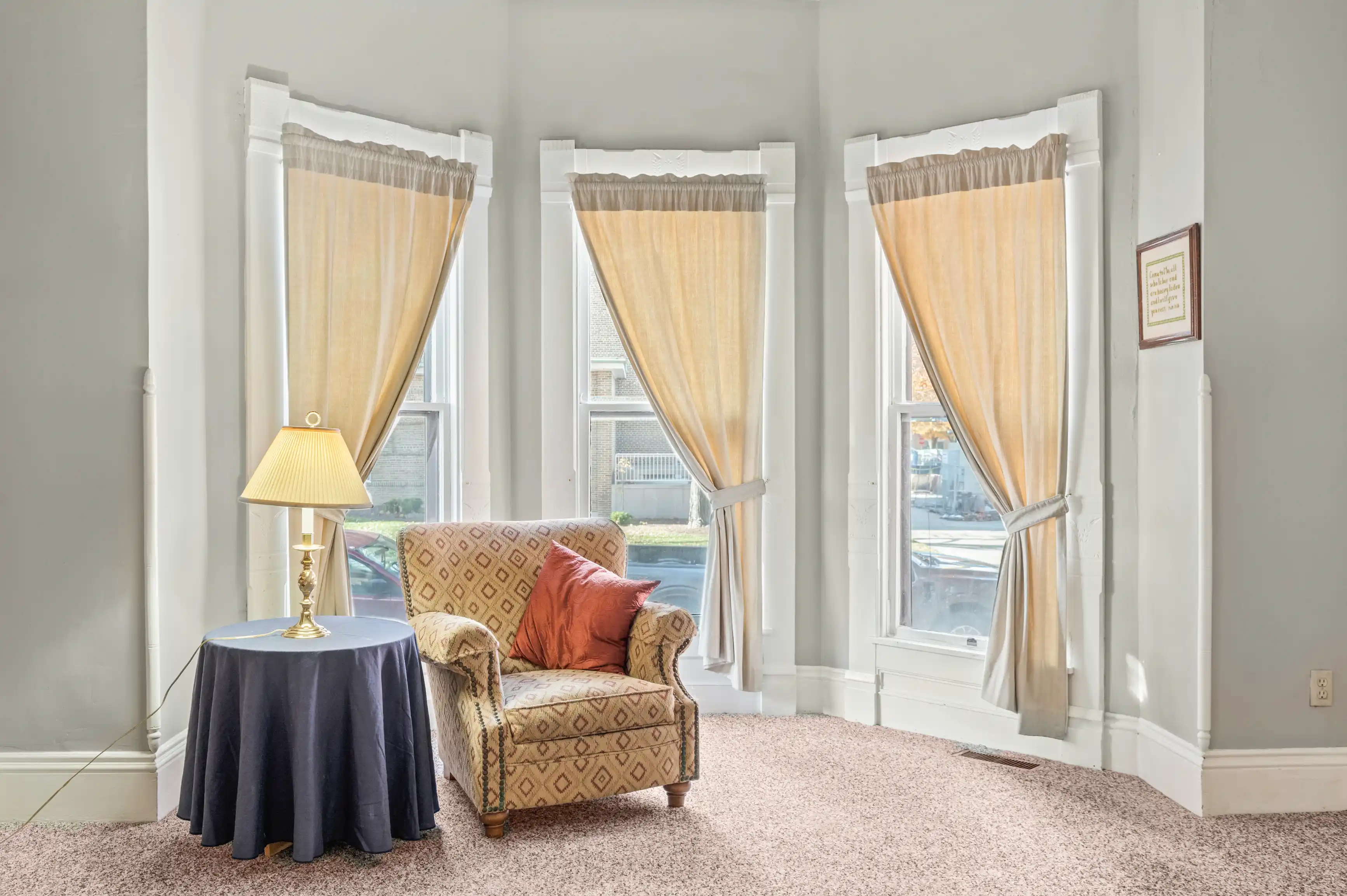 Cozy corner of a bright living room featuring elegant beige curtains framing a big window, a patterned armchair with a crimson pillow, and a classic table lamp on a round navy tablecloth.