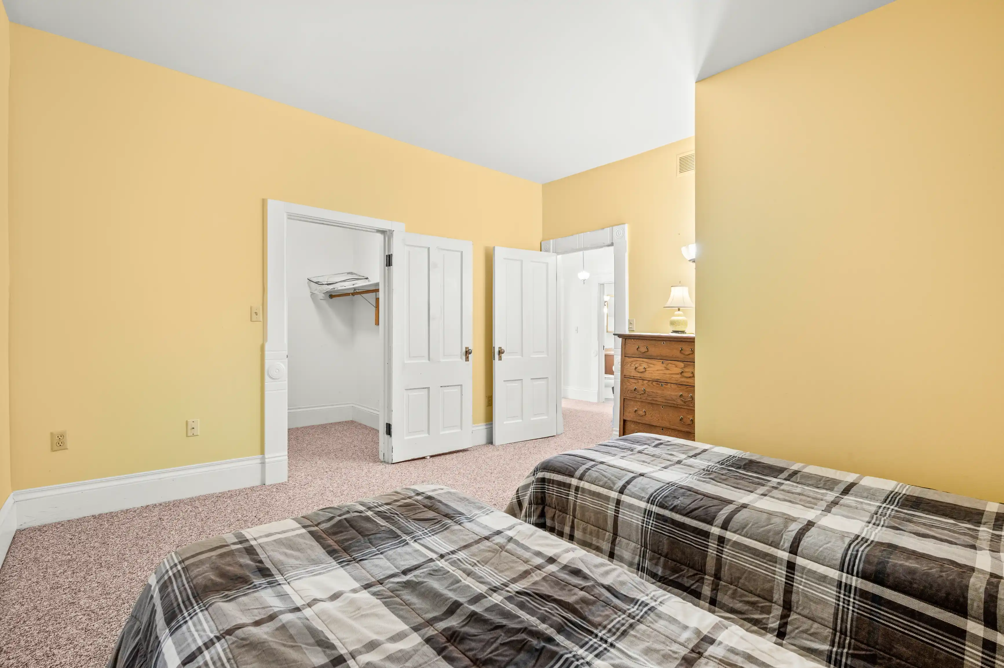 A well-lit bedroom with yellow walls, two plaid beds, an open closet, an ironing board, and wooden furniture.