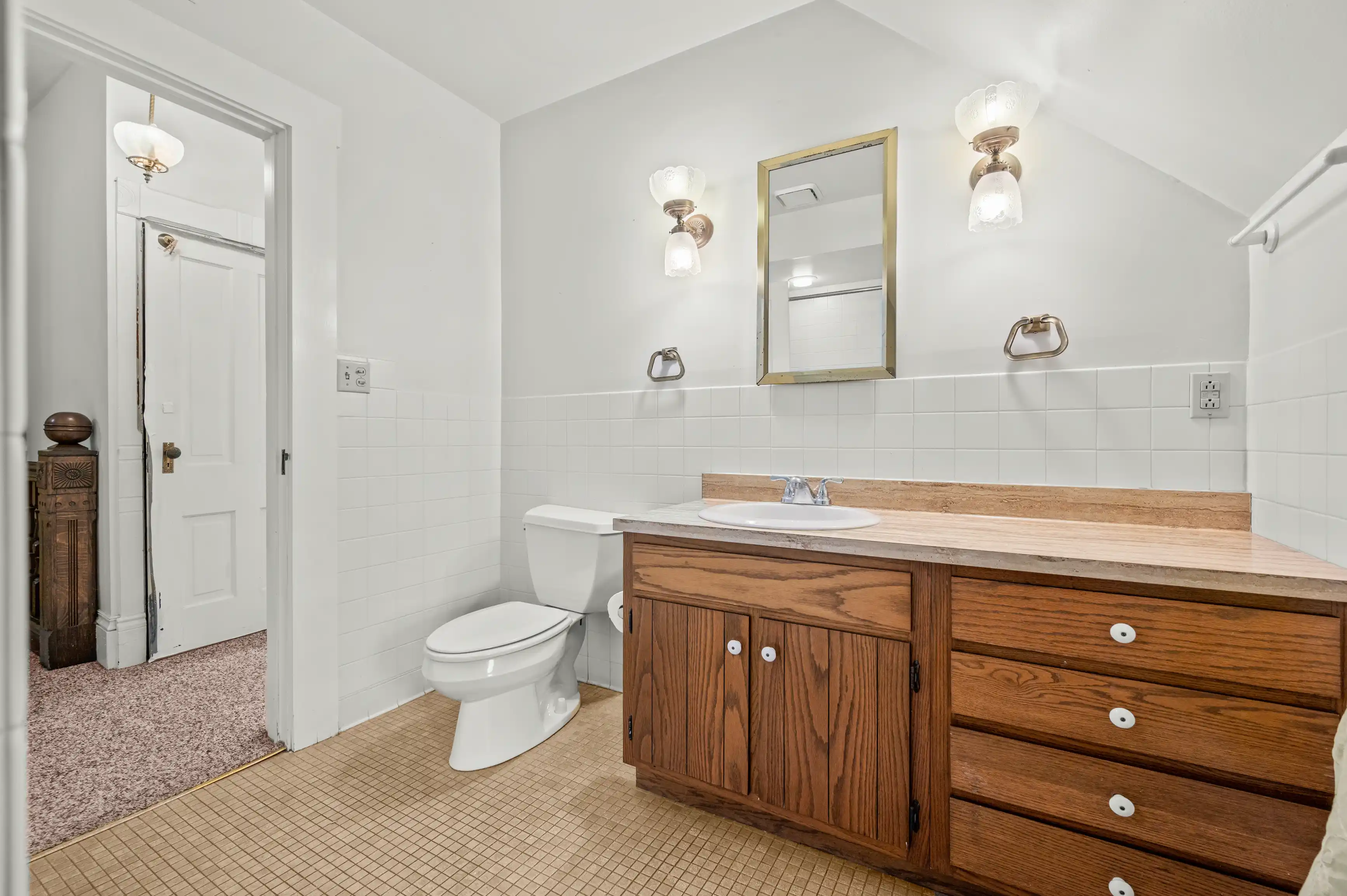 A bright and clean bathroom with white walls and tile, featuring a wooden vanity with white sink, wall mirror, toilet, tiled floor, and a light fixture above the mirror.