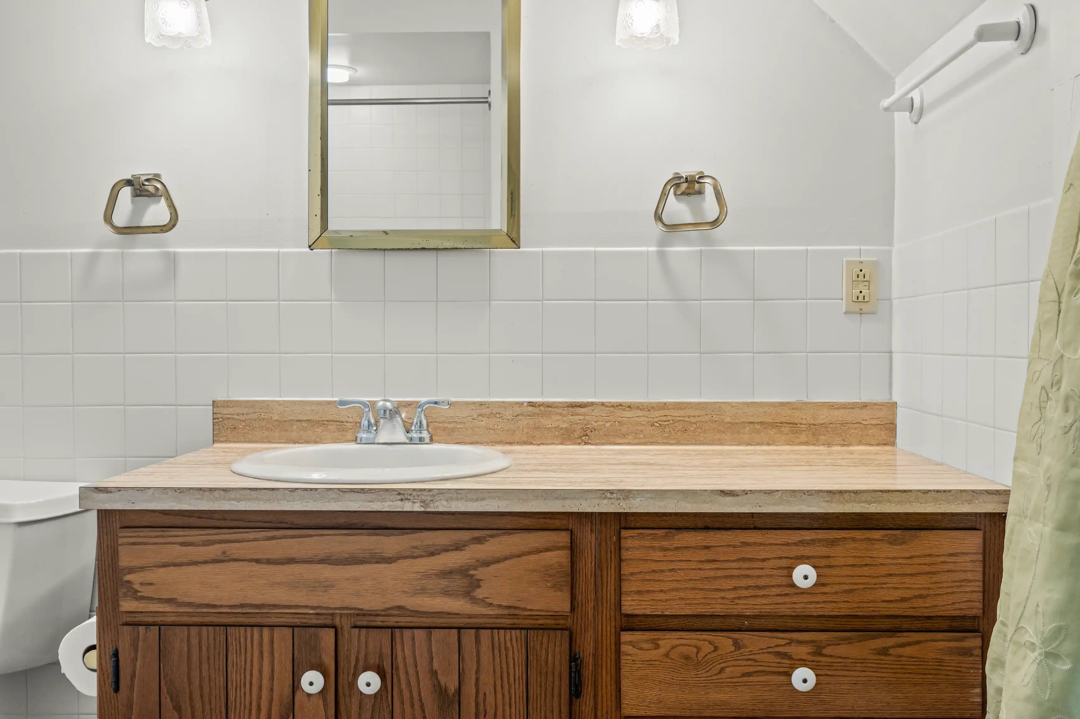 A neatly organized bathroom with white tiling, a wooden vanity with a beige countertop and white sink, a rectangular mirror with metallic frame above the sink flanked by wall-mounted lights, and a partial view of a bathtub with a pale green shower curtain to the right.