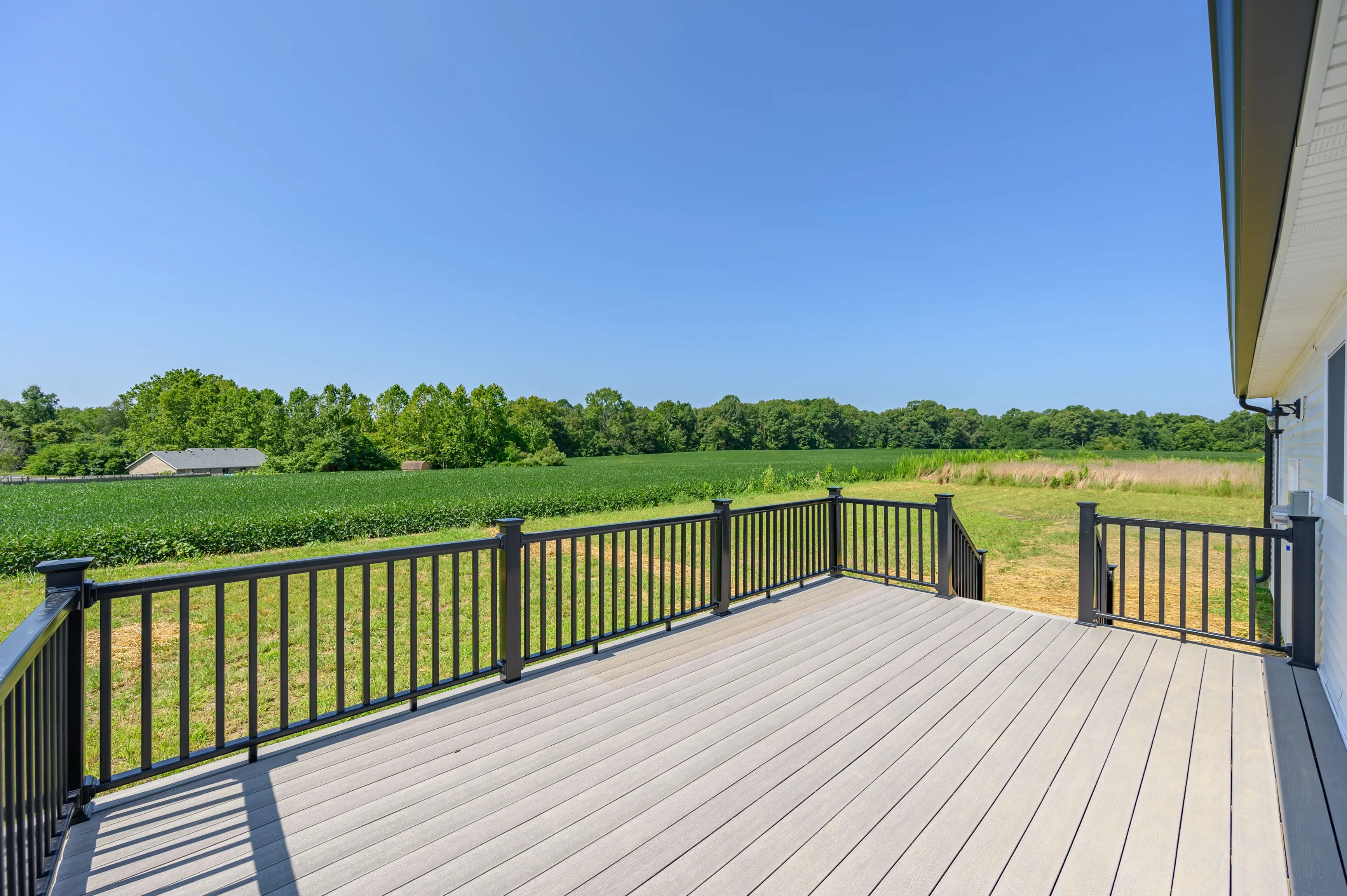 Spacious deck with railing overlooking a lush green field and trees under a clear blue sky.