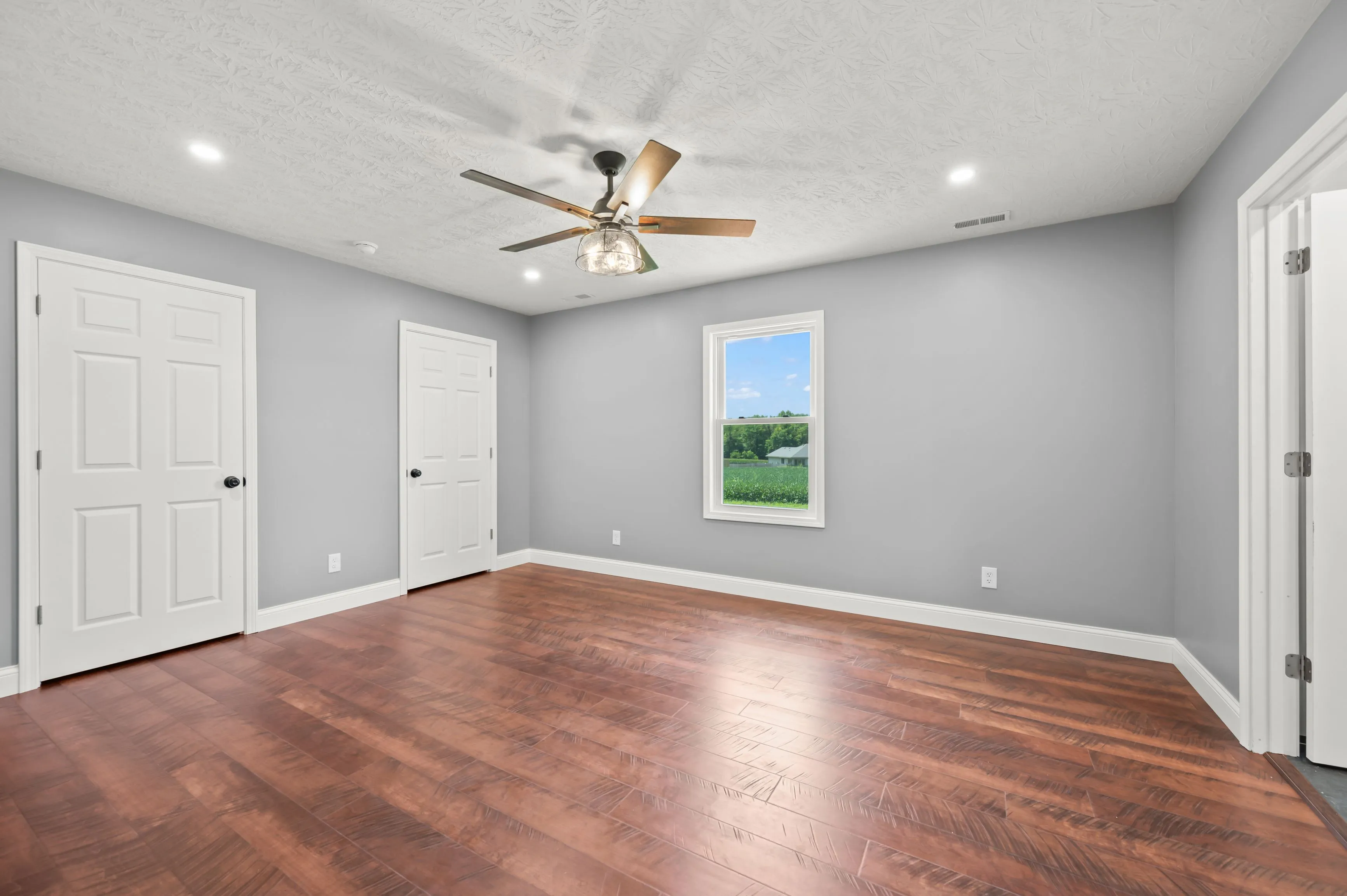 Bright empty room with gray walls, wooden floor, ceiling fan, recessed lighting, and a window with a view of greenery outside.