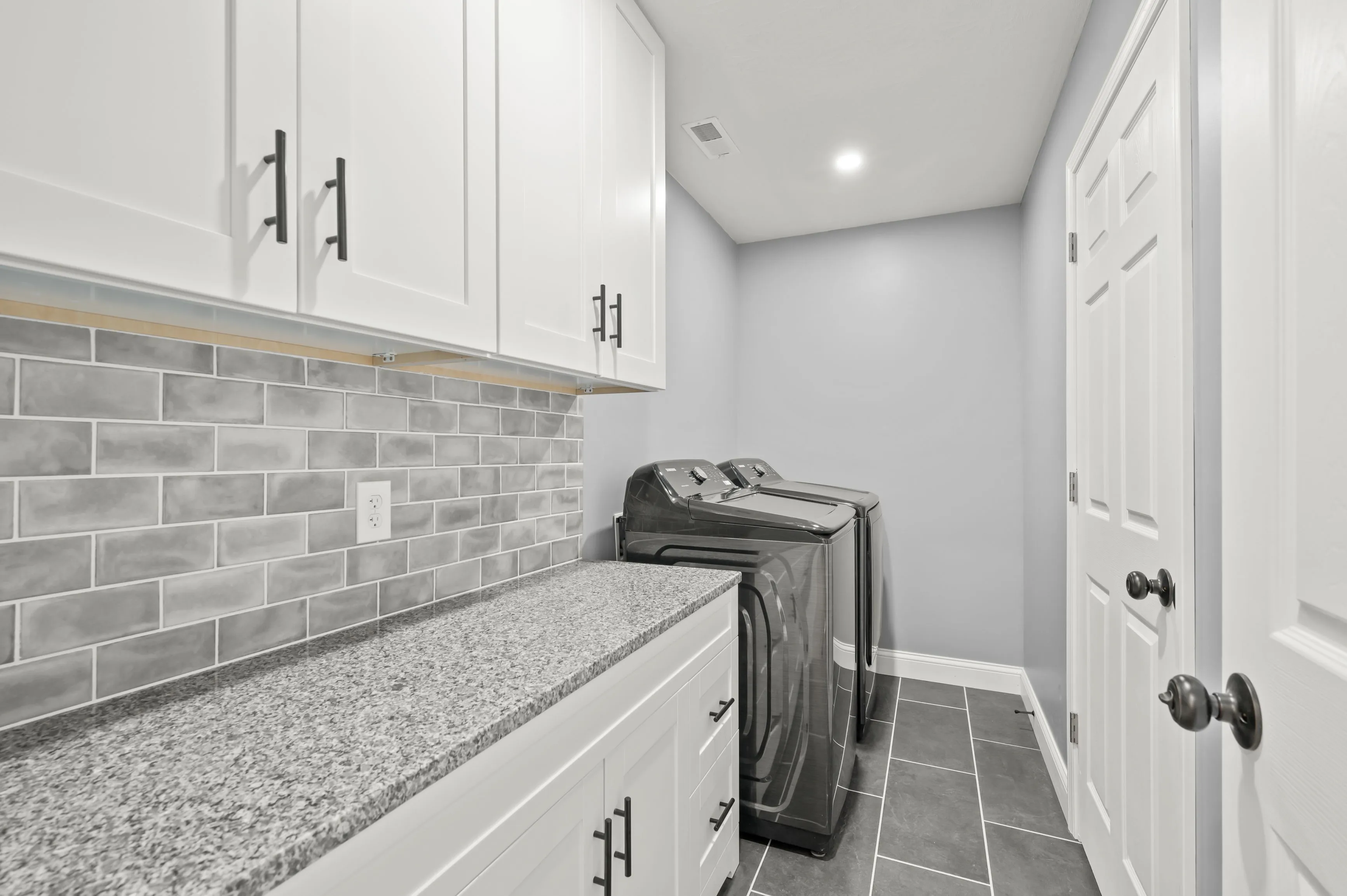 Modern laundry room with white cabinetry, gray granite countertops, gray subway tile backsplash, and black appliances.