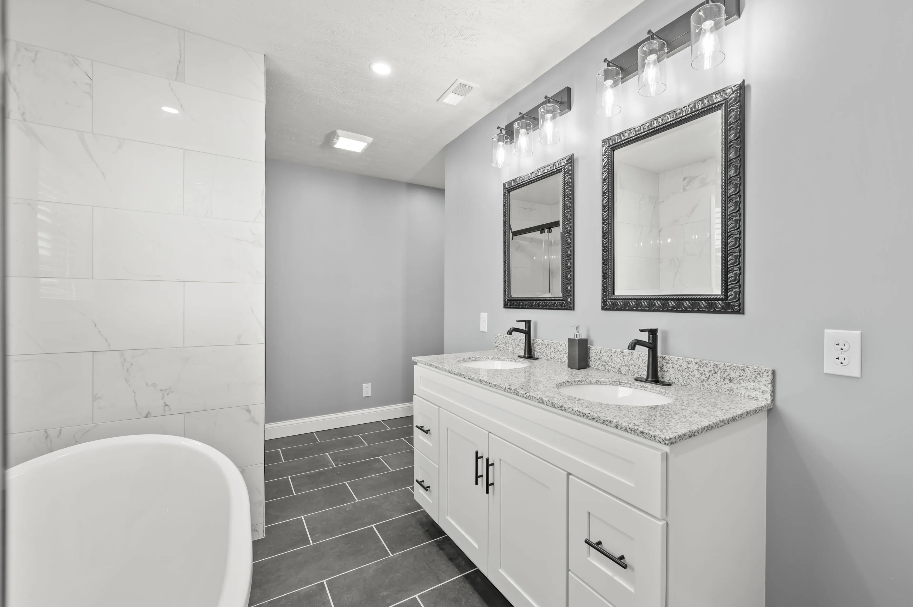 Modern bathroom interior with a white freestanding tub, double vanity with black faucets, matching framed mirrors, and gray tiled flooring.
