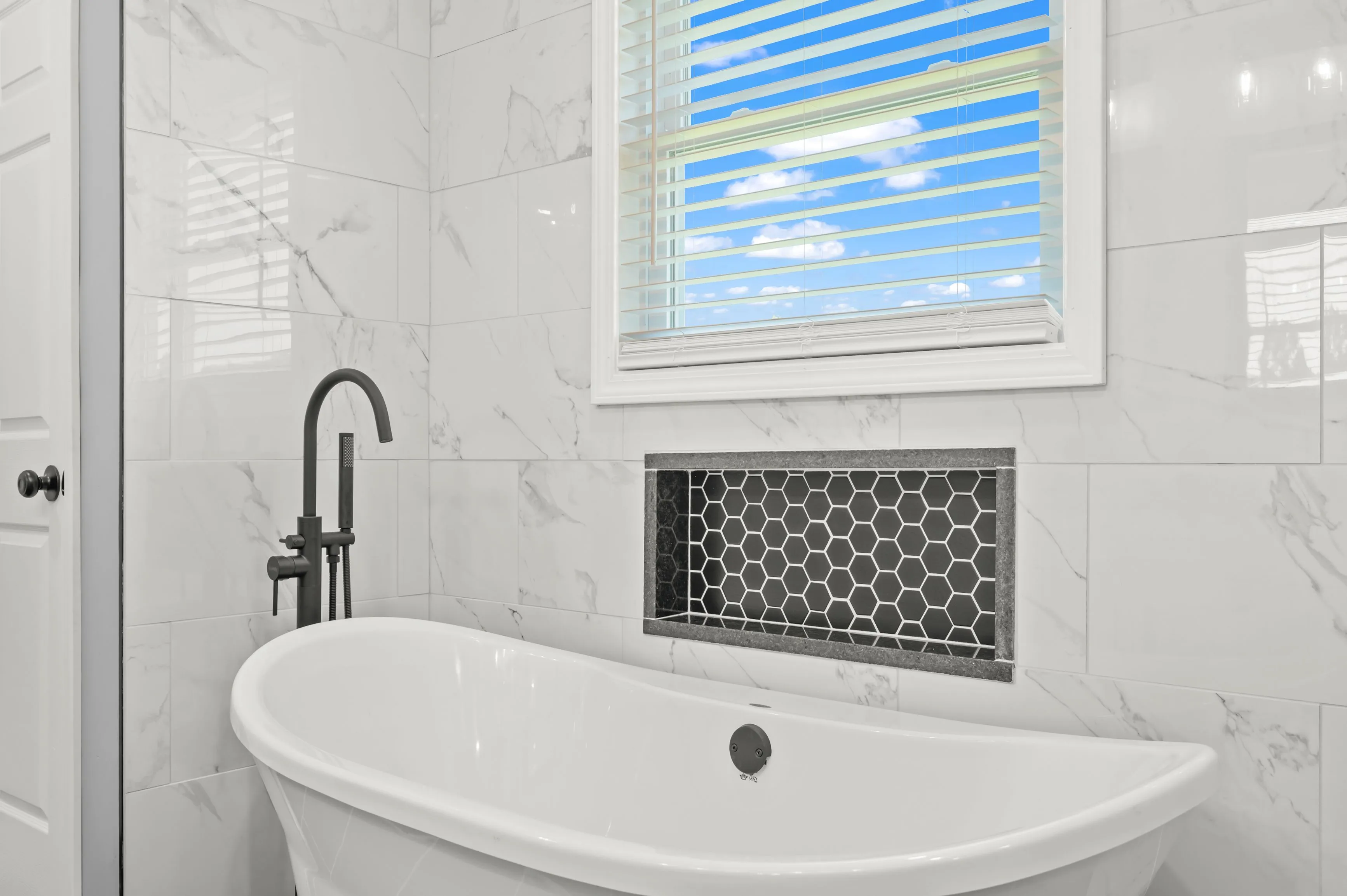 Modern bathroom interior with a white freestanding bathtub, a matte black faucet, white marble tiles, and a window with blinds partially showing a blue sky.