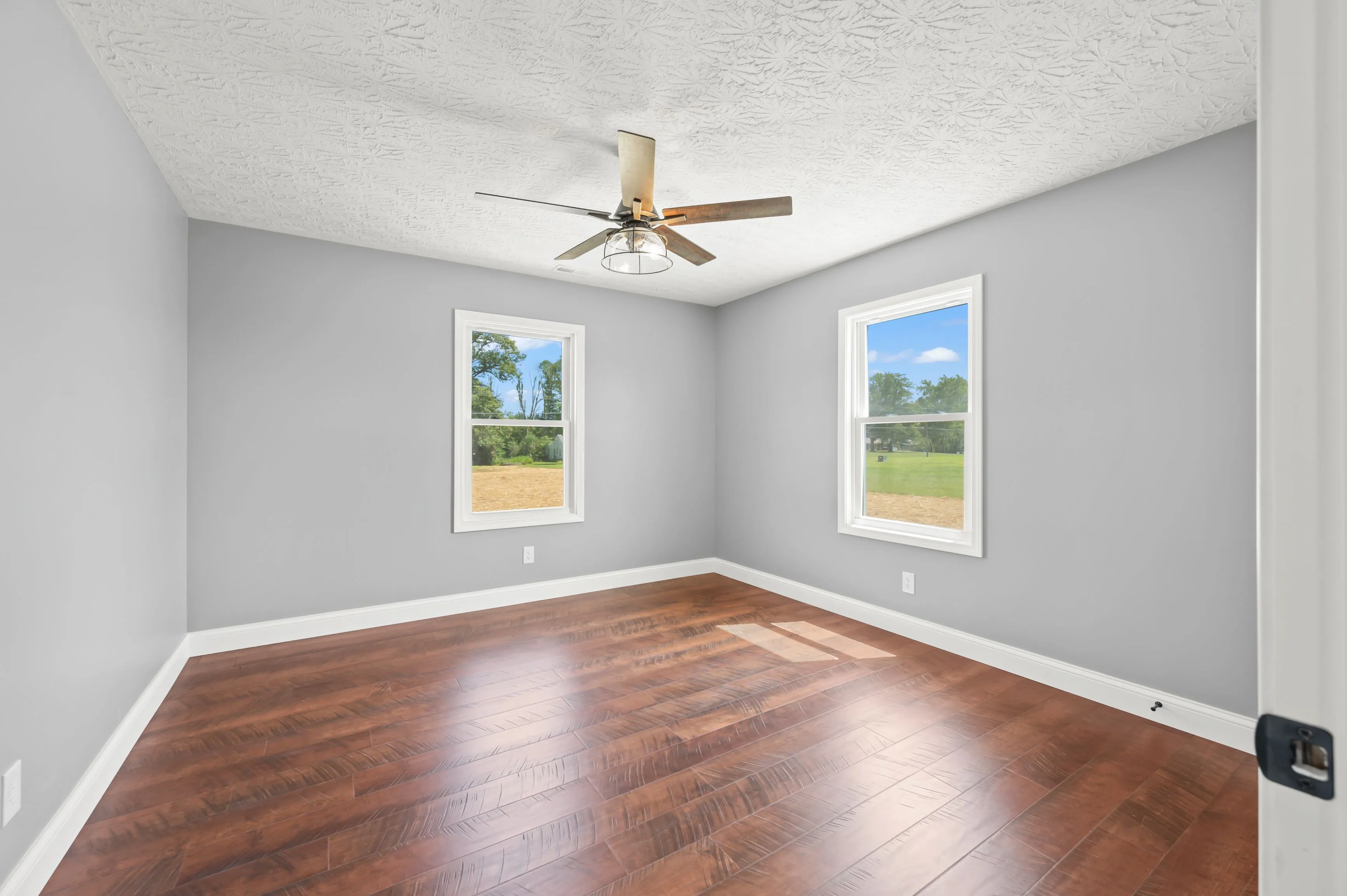 Empty room with gray walls, hardwood floors, a textured ceiling, and a ceiling fan, with two windows showing a view of trees and a lawn.