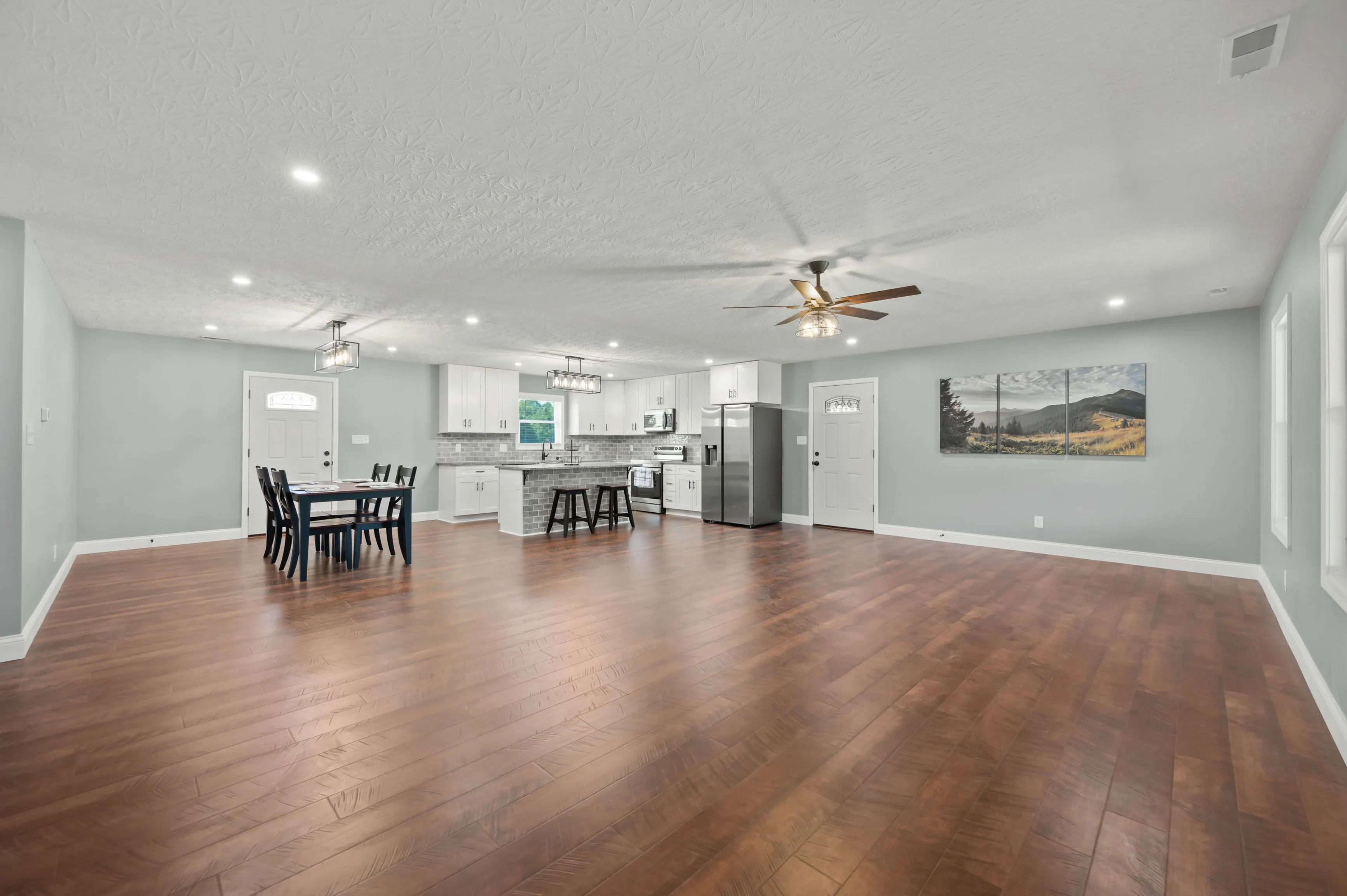 Spacious modern kitchen with white cabinetry and appliances connected to dining area with a dark wooden table and chairs, polished hardwood floors, overhead lighting, and a landscape painting on the wall.