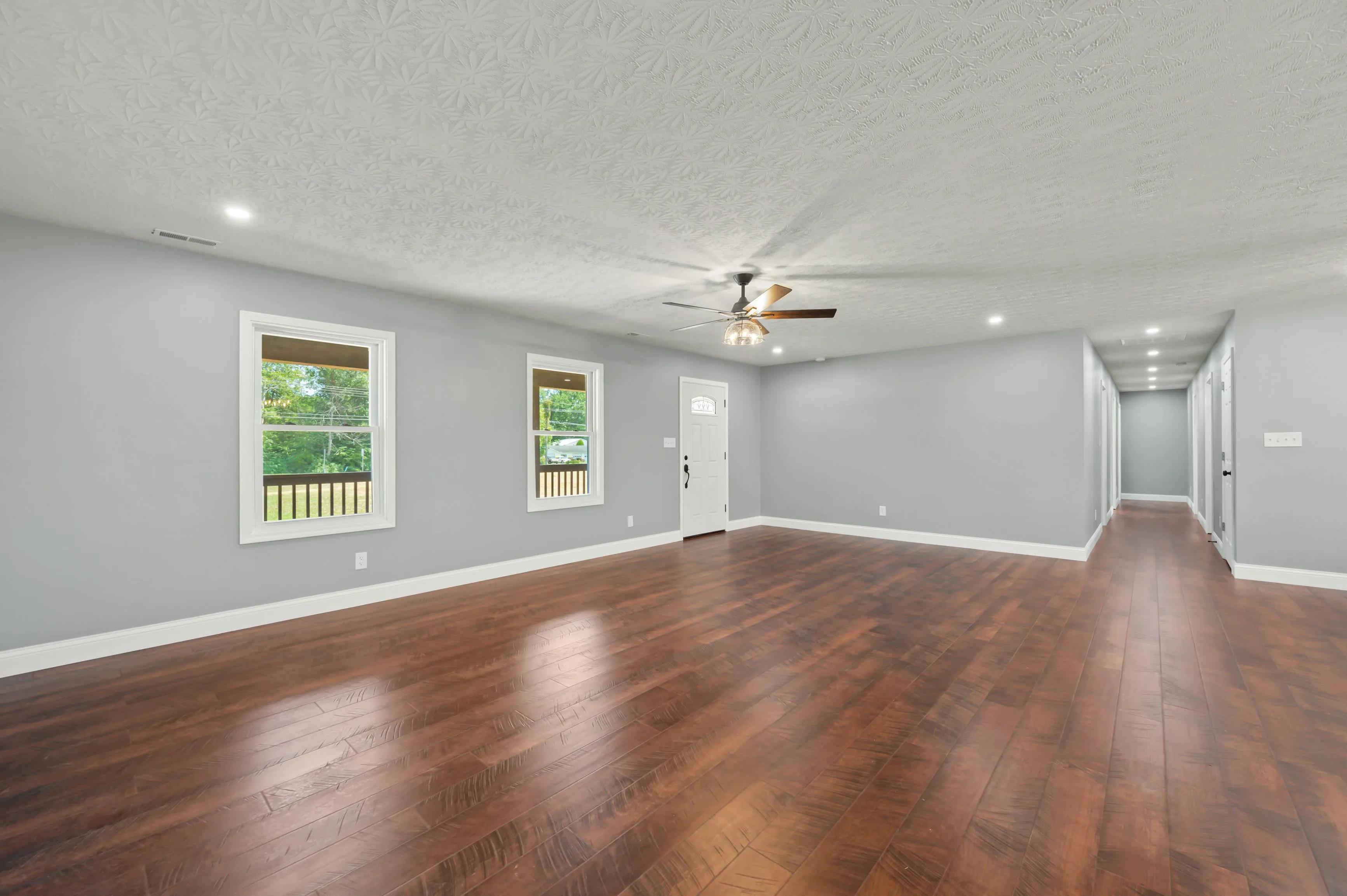 Empty room with polished wooden floor, light gray walls, white ceiling with textured pattern, ceiling fan, two windows and a door leading outside.