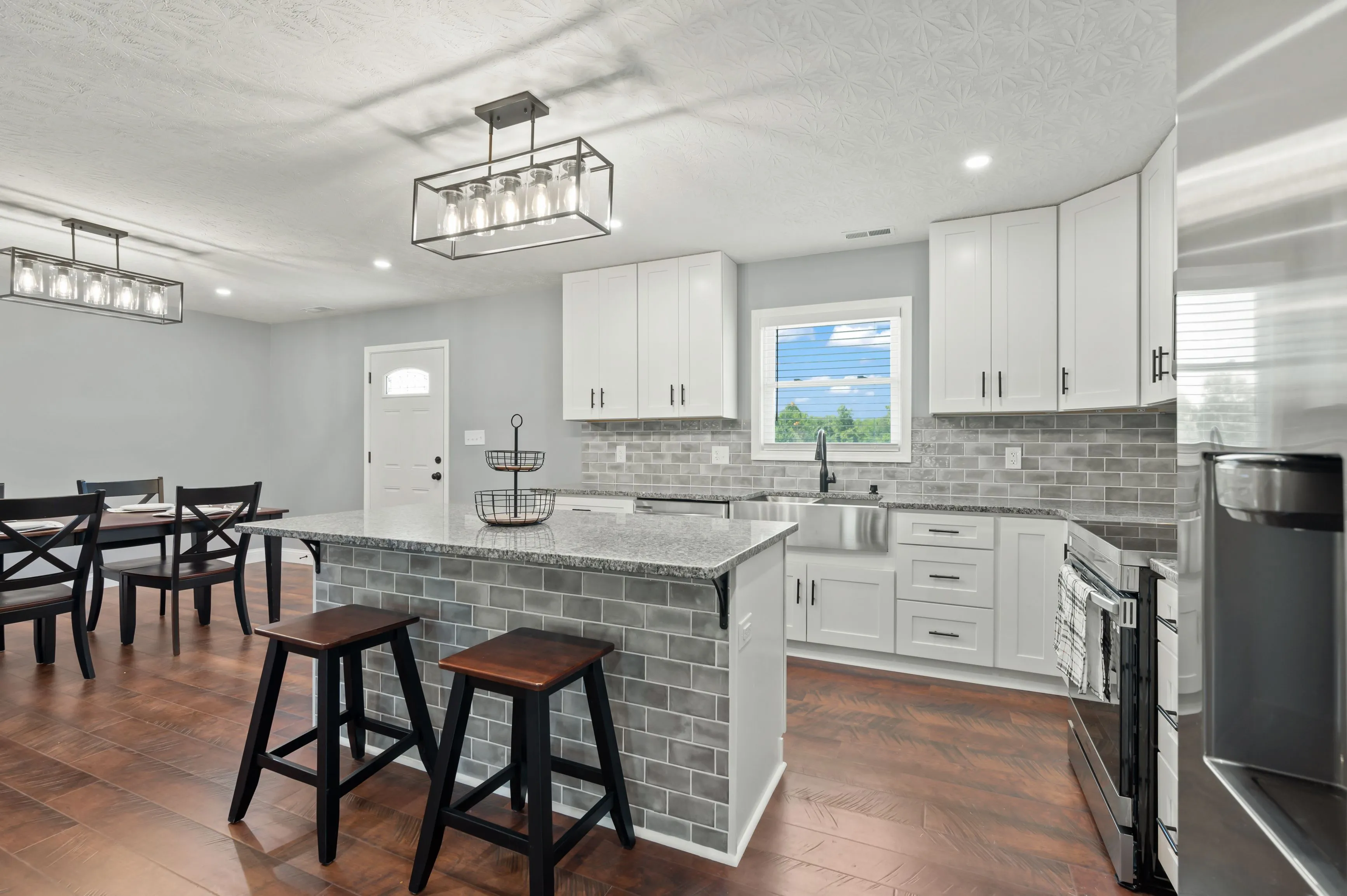 Modern kitchen interior with granite countertops, white cabinetry, stainless steel appliances, and a dining area with stylish lighting fixtures.